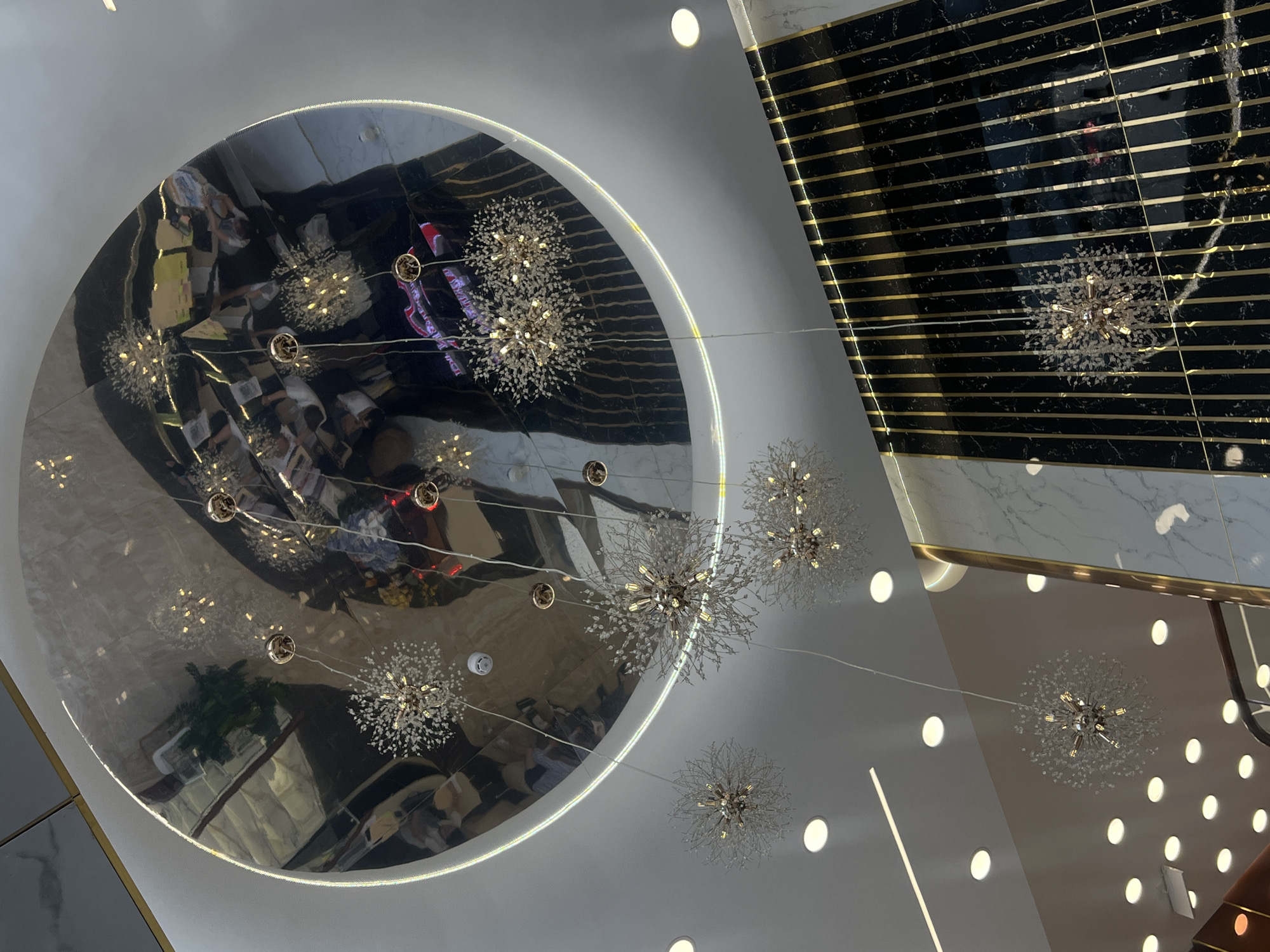 The image shows an upward view of the interior ceiling of a building. The ceiling features a large, circular, mirrored panel that reflects the details of the room and the lighting fixtures. Hanging below the mirrored panel and across the ceiling are several decorative light fixtures resembling dandelion heads, suspended by wires. The ceiling surface is white with evenly spaced recessed lights. An area of the ceiling adjacent to the mirrored panel features a black and gold striped design. The image's angle and the mirrored surfaces make it difficult to identify the location without further information. The location is in the city of Nha Trang, Vietnam.