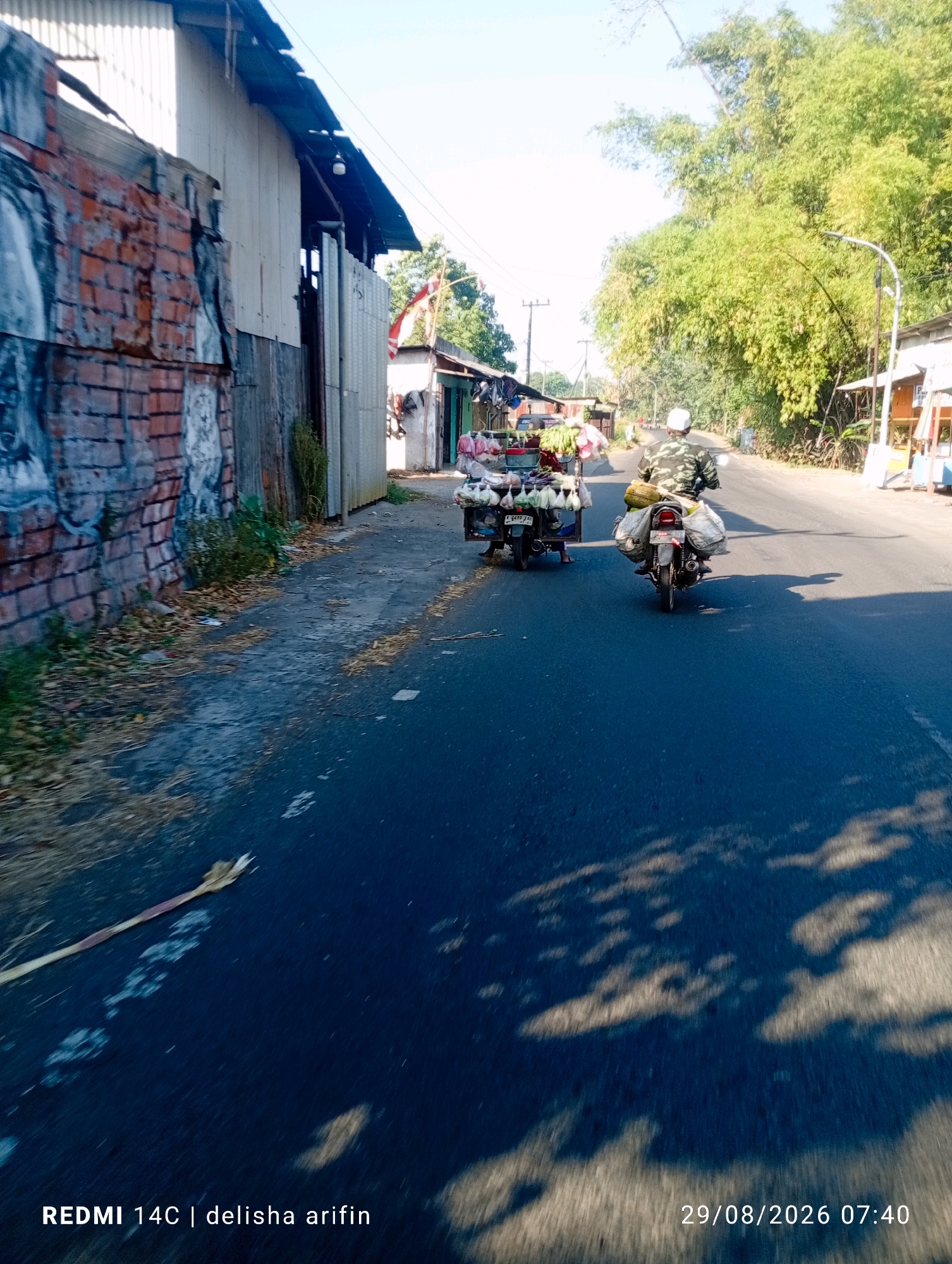 A street scene in Karang Asem, Indonesia, shows a paved road with a red brick wall on the left, covered in peeling paint and graffiti. A corrugated metal building with a tin roof is attached to the wall. Further down the road, a vendor's cart laden with produce, including bananas, is pulled by a motorcycle. A second motorcycle, also carrying sacks and a durian fruit, is in front of the vendor's cart. Both motorcycles are traveling away from the viewer. The road is lined with trees and some small roadside structures. The sky is bright and sunny, casting shadows on the road. The bottom right corner of the image displays a timestamp: "29/08/2026 07:40" and text that reads "REDMI 14C | delisha arifin".