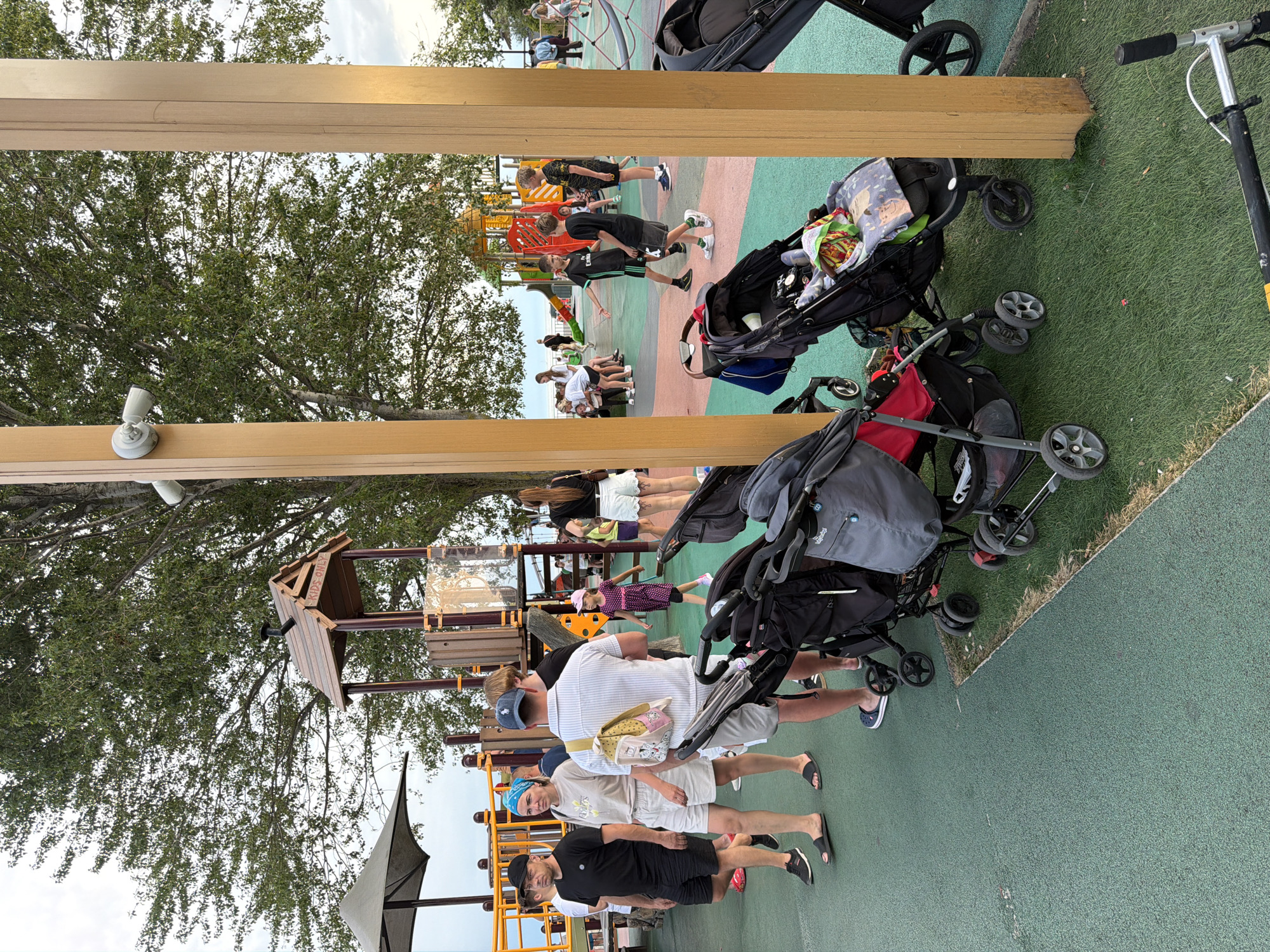 This is a photograph of a playground in Sochi, Russia. The playground appears to be a public space, possibly in a park or near a beach, given the presence of trees and what looks like a view of water in the background.
In the foreground, there are several strollers, indicating that many families with young children are present. The strollers are various brands and colors, some are empty and some appear to have items inside them.
To the left of the frame, a group of adults are standing and walking. One man in a black t-shirt and shorts is looking to his left. Beside him, a woman with a blue headscarf and a light-colored t-shirt and shorts is also looking to her left. Another man in a striped shirt and shorts with a backpack is facing away from the camera.
In the middle ground of the image, there is a children's playground structure with colorful slides and climbing equipment. Several children are visible on and around the playground. A young girl in a pink polka-dot dress is walking away from the camera. Two boys in black t-shirts are interacting on the playground.
The playground surface is a mix of green artificial turf and a softer, colored material. In the background, more people can be seen enjoying the playground and the surrounding area. The sky is overcast, suggesting it might be late afternoon or evening. The overall scene depicts a lively and active family-friendly environment.