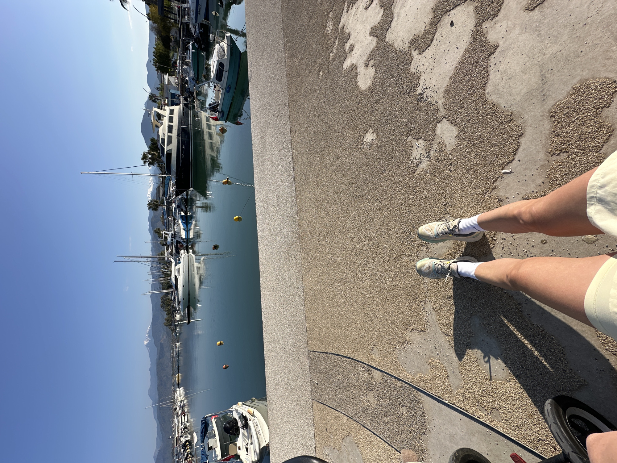 The image shows a person standing on a pier in Fethiye, Turkey. The harbor is filled with boats, and the mountains are visible in the background. The person is wearing sneakers and shorts. The pier is made of concrete, and there are some small pebbles on the ground.