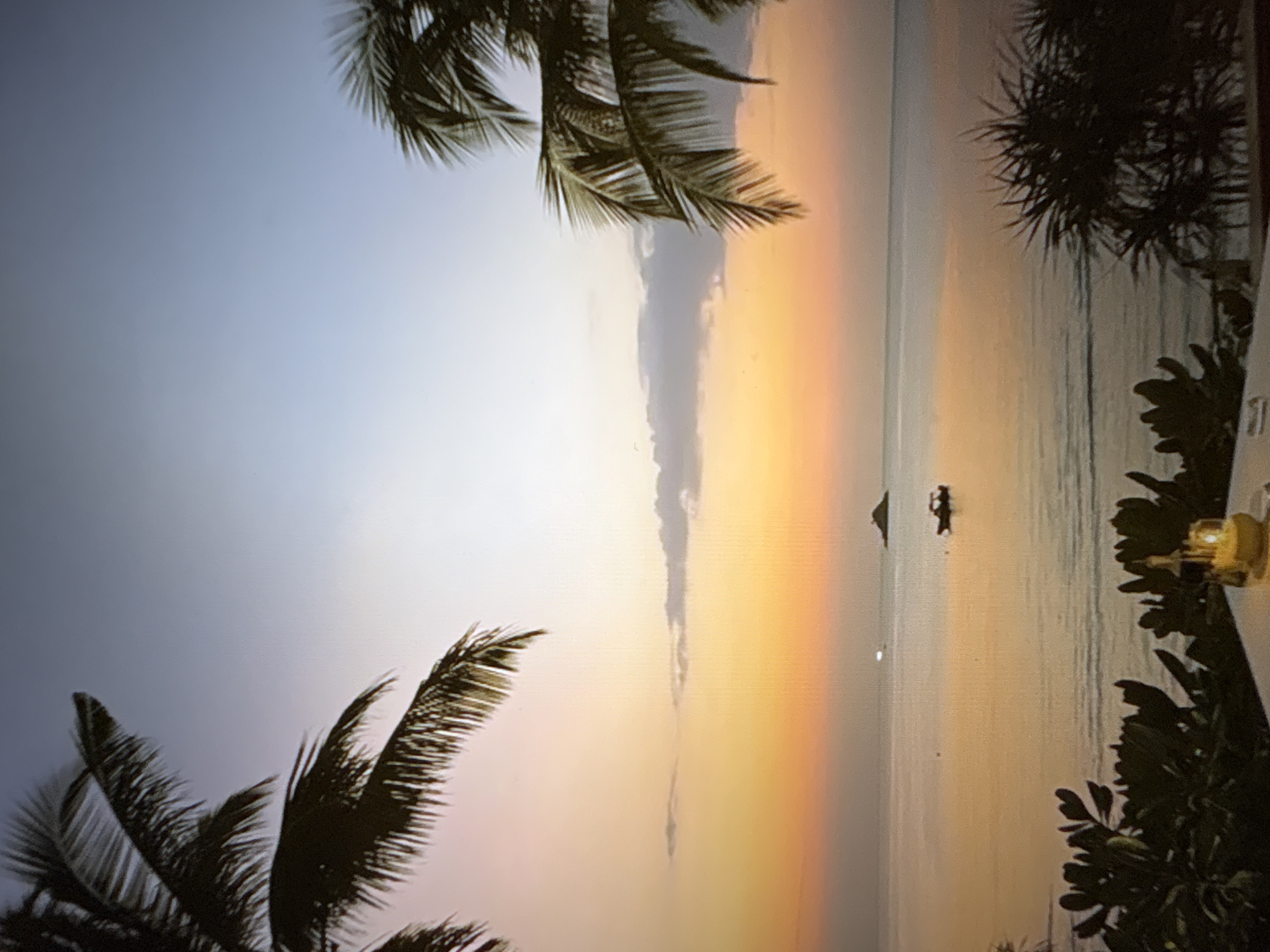The image depicts a serene ocean sunset viewed from a tropical location, likely a resort or restaurant. In the foreground, silhouetted palm fronds frame the scene from the top left and right. Below, lush dark green foliage is visible, partially obscuring a white table in the bottom right corner. On the table sits a lit candle in a yellow holder, casting a warm glow.
The middle ground features a calm body of water, reflecting the soft hues of the sunset. A small dark boat is visible in the middle distance, and a tiny island or landmass can be seen further out. Distant lights twinkle on the water.
The sky transitions from a soft gradient of pastel pinks and oranges near the horizon to a pale blue at the top. A few wispy clouds are scattered across the sky.
The scene evokes a peaceful, tranquil atmosphere, characteristic of a tropical evening. The image does not depict the city of Pori, Finland.