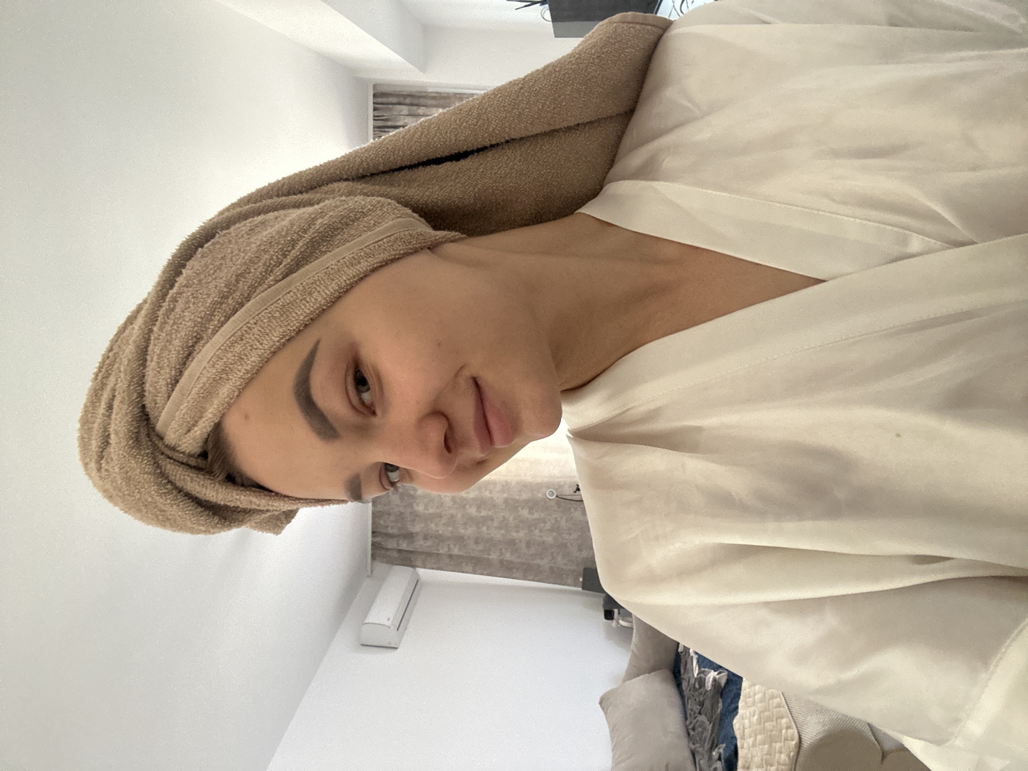A woman is depicted indoors, wearing a white robe and a brown towel wrapped around her head. An air conditioning unit can be seen on the wall. The room's interior also features a couch and curtains.