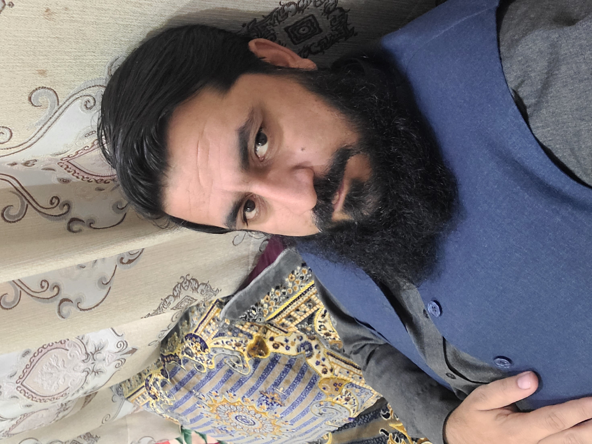 A man with dark hair and a full beard is reclining against a patterned cushion or pillow on what appears to be a sofa or bed. He is wearing a gray shirt with a blue vest or jacket. The backdrop consists of a beige fabric with scrolling, ornate patterns. The environment appears to be an interior space. This picture was taken in the city of Quetta City Tehsil, Pakistan.