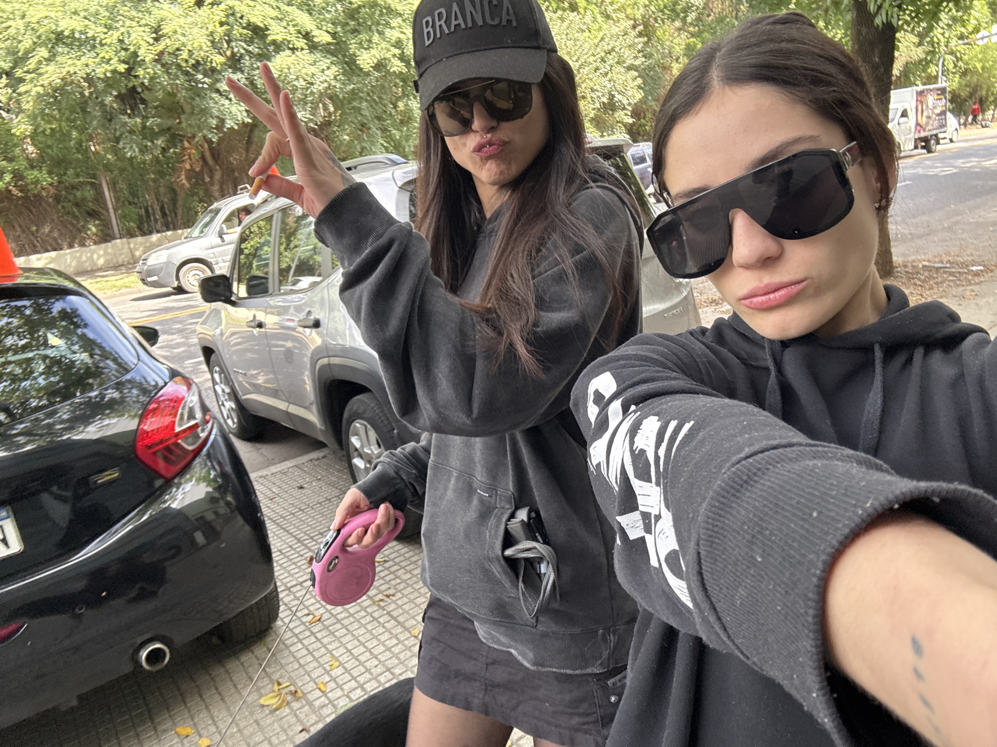 Two young women are posing for a selfie on a city street in Buenos Aires, Argentina. The woman on the left is wearing a black baseball cap with the word "BRANCA" on it, sunglasses, and a grey hoodie with dark-colored shorts. She is holding a pink retractable dog leash in her hand and making a hand gesture with a cigarette between her fingers. The woman on the right is wearing sunglasses and a black hoodie. Cars are parked along the street, and trees are visible in the background. A delivery truck is seen down the street to the right.