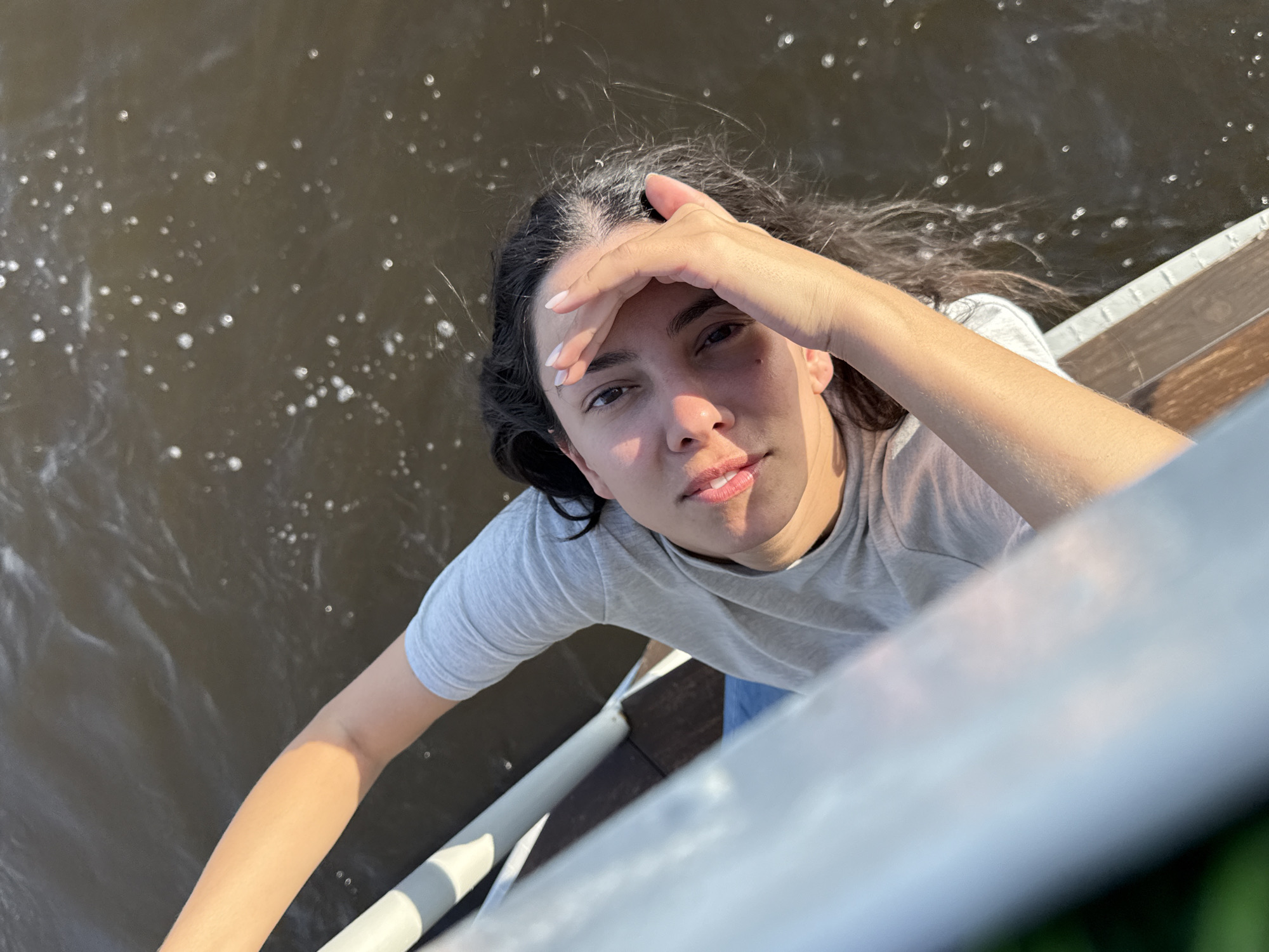 A young woman with dark hair is captured in a high-angle shot, leaning over a railing that appears to be part of a pier or boardwalk. She is wearing a light grey t-shirt and blue jeans. Her right hand is raised to her forehead, shielding her eyes from the sun. The water in the background is murky and reflects sunlight, creating small, bright circles on its surface. The railing in the foreground is white and blue, with wooden planks visible beneath it. This scene is likely located in Chaikovsky, Russia, possibly near the Kama River or a similar body of water, given the city's geography.