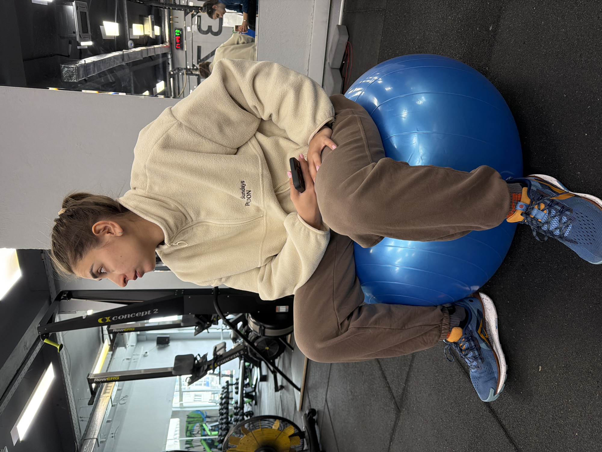 A woman is seated on a blue exercise ball, holding a black cell phone, likely in a gymnasium, potentially in Sofia, Bulgaria. She is wearing a cream-colored fleece jacket with the word "SUNDAYS" vertically stitched on the left chest area, along with brown pants and blue athletic shoes with orange accents. The gymnasium contains Concept 2 rowing machines and dumbbell racks in the background. A mirror reflects the gym's interior, including a person visible in the reflection. The floor is made of dark rubber material.