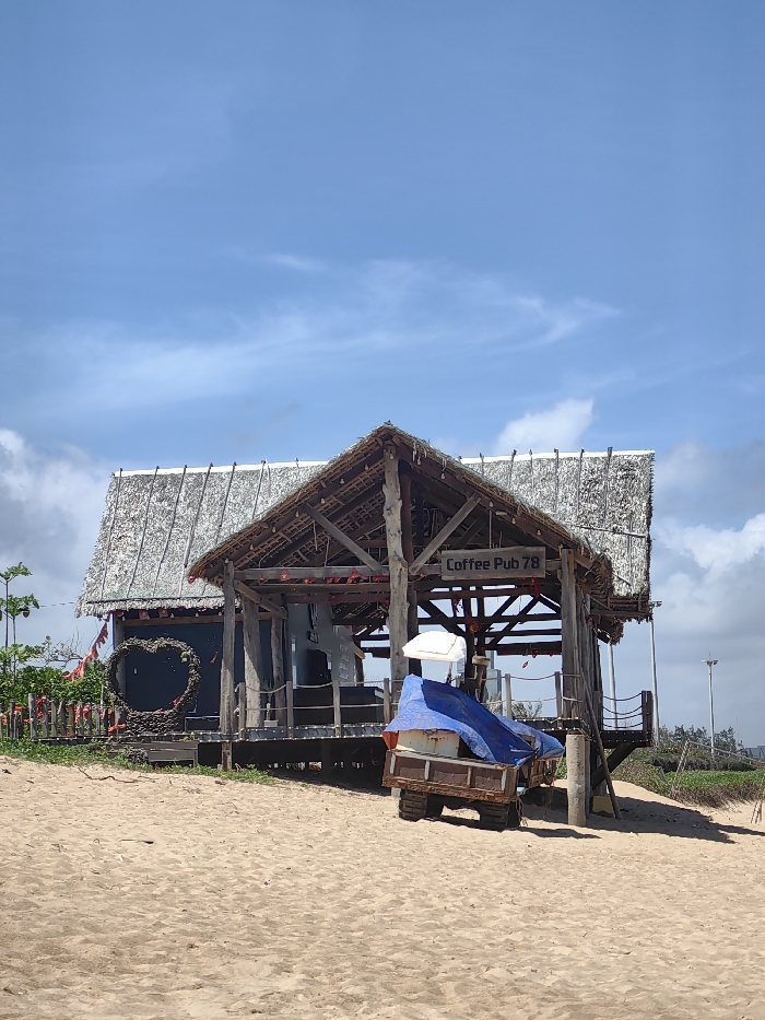 This image depicts a beach scene in Tuy Hòa, Vietnam, under a bright blue sky with scattered white clouds. In the midground, two rustic structures stand on sandy ground. To the left, a dark-colored building with a steeply pitched thatched roof features a large, decorative heart-shaped frame constructed from natural materials, situated on its exterior wall. A line of small red heart-shaped ornaments or flags extends to the left of this building, leading towards green vegetation. To the right of the first structure is an open-air pavilion or hut, also with a thatched roof supported by exposed wooden beams and posts. A wooden sign reading "Coffee Pub 78" is affixed to one of its overhead beams. The pavilion has a raised wooden deck with rope railings. Positioned on the sand underneath the open-air pavilion, a small, multi-wheeled, brown, metal-framed cart is visible, largely covered by a blue tarp. A folded white beach umbrella is partially visible next to the tarp on the cart. No individuals are present. Sparse green vegetation and a utility pole are visible in the far background to the right. The foreground consists entirely of light-colored sand.