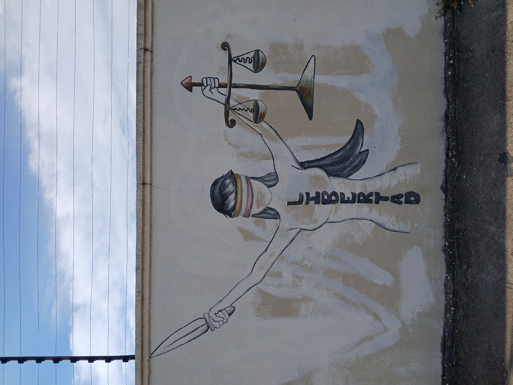An outdoor mural painted on a light-colored wall is the central subject of the image, set against a blue sky with power lines on the left and a concrete structure on the right. The mural depicts a stylized female figure, possibly representing a form of Lady Justice or Liberty.
The figure has a blindfold composed of yellow and pink/red bands. Her left arm extends downwards, clutching a dagger with a pointed blade and a dark handle. Her right arm is raised, holding aloft a set of scales of justice. These scales are depicted with a stylized, arrow-like shaft, and one of the scale pans appears broken or unusually shaped, while the other is intact. Dark, stylized wings are visible on her back. Vertically written in black capital letters on her torso is the Spanish word "LIBERTAD" (Freedom). The background of the mural itself is a mix of light tan/beige and white brushstrokes, suggesting a painted backdrop.
The surrounding environment includes a bright blue sky with scattered white clouds, traversed by numerous thin, parallel power lines in the upper left portion of the image. Along the right side, a weathered concrete curb or wall runs vertically, showing cracks and some small green plant growth. The overall impression is one of a public art piece in Ciudad Guayana, Venezuela, conveying themes of justice and freedom, possibly in a state of struggle or transformation. The image appears to be taken in daylight, given the clear sky.
