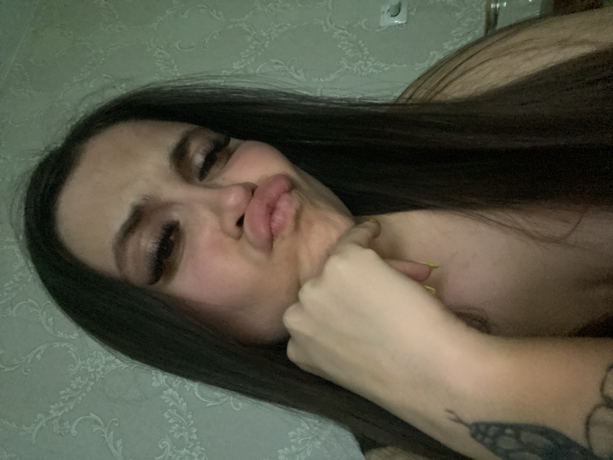 A woman with long, dark brown hair is lying down, puckering her lips for a kiss. She is resting her chin on her right hand, and her eyes are closed. Her left arm is visible, and there appears to be a tattoo on her forearm. The background is a light green patterned wall. The location is possibly Chaikovsky, Russia.