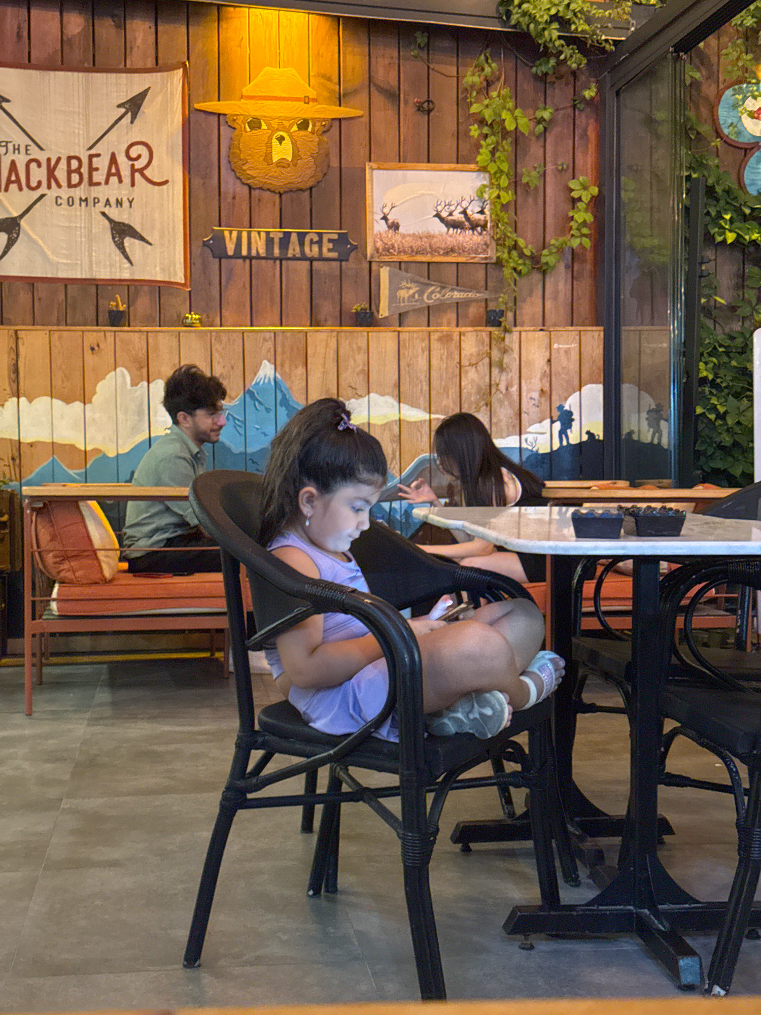 In Konyaaltı, Turkey, a child sits at a table, engrossed in a mobile phone, positioned in the foreground. The child is seated in a black chair, wearing a lavender top and shorts. To the left, a young man in a green shirt is seated on a bench with orange cushions. Behind him, a wall with a painted mountain and cloud mural stretches across the background. The wall is adorned with various decorative items, including a "VINTAGE" sign, a bear mascot, and a framed picture of deer. A lush green vine hangs from above, adding a touch of nature to the scene. The room is furnished with dark tables and chairs, and the floor is tiled in a neutral tone.