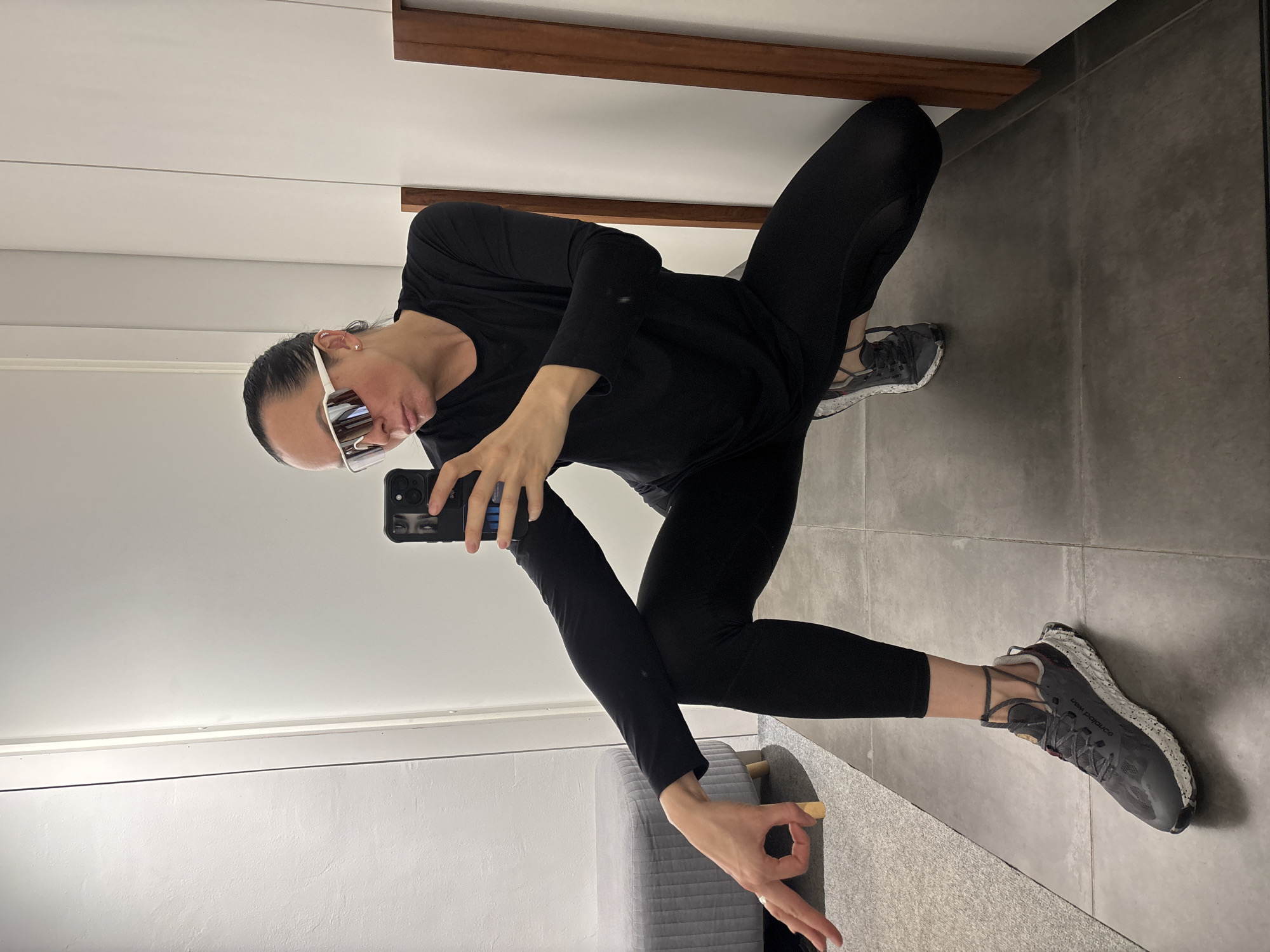 A woman in black athletic clothing and white-framed sunglasses is taking a selfie in a bathroom. She is squatting and holding her phone up, pointing the camera towards herself. The phone has a black case with an image of eyes on it. The woman is making an "okay" gesture with her left hand. She is wearing gray athletic shoes with a speckled pattern. The bathroom has a white wall with a wooden accent at the top, a gray tiled floor, and a gray upholstered chair or bench partially visible in the lower left corner. The location is presumably indoors. There is no evidence to indicate that the image was taken in Chaikovsky, Russia.