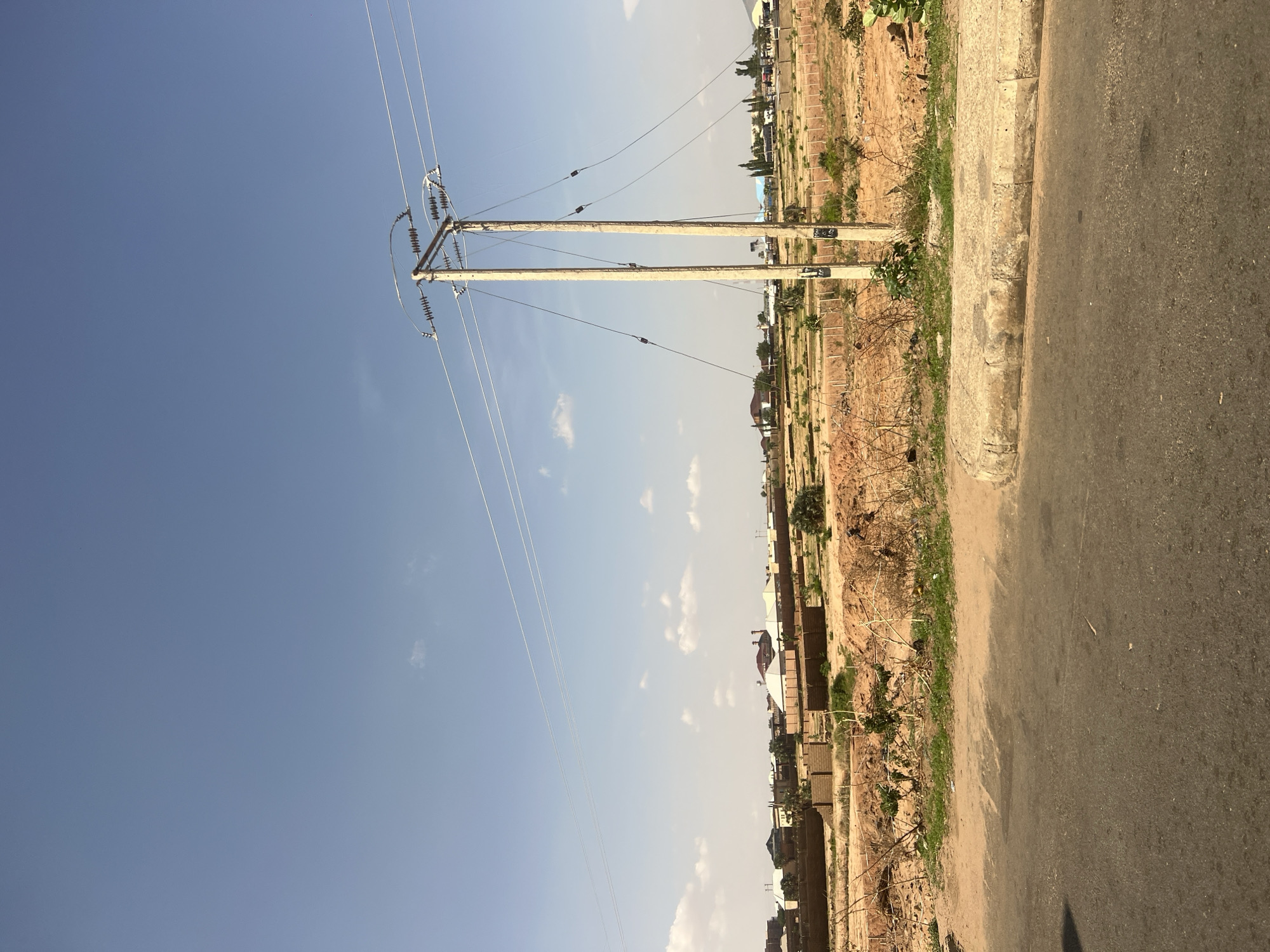 This image depicts an outdoor environment in Gombe, Nigeria, under a clear, light blue sky featuring scattered cumulus clouds. No people are visible within the frame.
The foreground is occupied by a dark asphalt road surface on the left, which transitions into a concrete curb on the right, separating it from an unpaved area.
The mid-ground consists of reddish-brown, dry earth with sparse, desiccated vegetation, including patches of green grass and bare, twiggy bushes. Prominently positioned on the right are two light grey concrete utility poles, standing vertically and parallel to each other. These poles support multiple horizontal power lines and associated insulators and connecting wires. Further into the mid-ground, multiple rows of partially constructed low walls or foundations made of concrete blocks are visible across the undeveloped land, indicating ongoing or planned construction. Several mounds of excavated reddish-brown soil are also present.
In the background, a cluster of residential or commercial buildings with varying roof designs and colors (some flat, some pitched, some red, some light-colored) is discernible, suggesting a developed settlement in the distance. Additional sparse trees and distant topographical features are faintly visible on the horizon. The overall lighting suggests natural daylight, with a dry environmental condition.