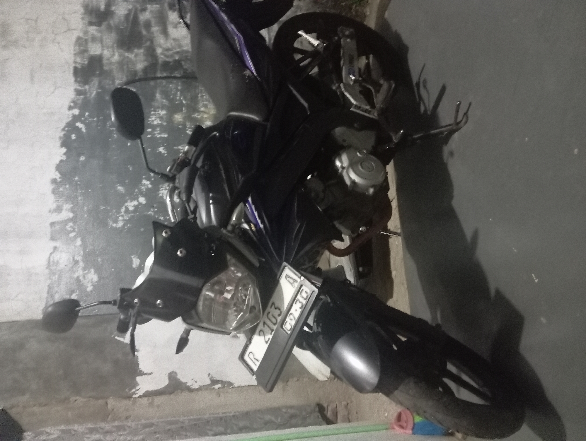 A dark-colored motorcycle is parked on a gray, uneven surface, casting a shadow to the right. The motorcycle is angled towards the viewer's left, with its front wheel slightly turned. The license plate, affixed to the front of the motorcycle, reads "R 2103 A" and below that "09.3G". The background consists of a textured, light-colored wall with dark smudges and cracks. A green object, possibly a broom or a pole, is visible on the far left side of the image. The lighting suggests an indoor or dimly lit outdoor environment. This scene is located in Kutasari, Indonesia.
