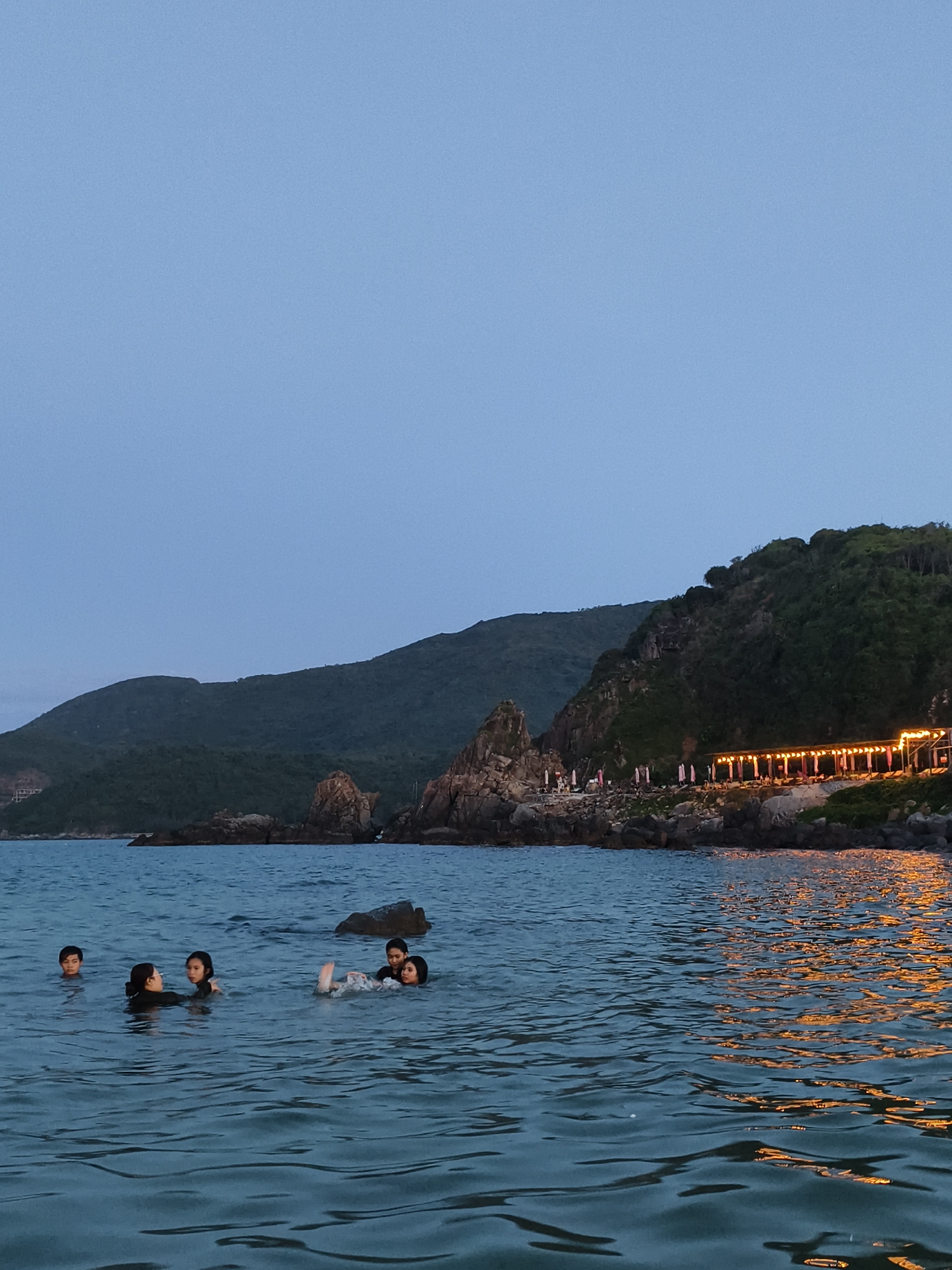 In Vĩnh Lương, Vietnam, the image captures a serene coastal scene at dusk. Five individuals are immersed in the calm, dark blue water, their bodies partially submerged. In the foreground, on the left, a group of three appears to be interacting, with two facing each other and one looking towards the viewer. To the right, another individual lies on their back, legs up, while a fifth person is visible further to the left. The water surface is gently rippled, reflecting the warm, yellow lights of a beachfront establishment situated on a rocky incline in the background. This establishment features a long, covered structure with multiple lights illuminating the area. The shoreline is characterized by dark rocks and lush green vegetation climbing up the steep, mountainous terrain. The sky above is a clear, pale blue, indicative of twilight.