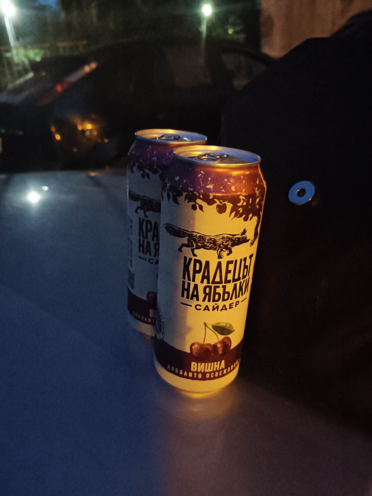 The image shows two cans of cider placed on a dark, reflective surface, possibly the hood of a car. The cans are light-colored with red and brown designs. The can in the foreground has text that appears to be in Cyrillic script, along with an illustration of a fox and cherries. The background is dark and out of focus, with some blurred lights that suggest an outdoor, possibly nighttime setting. There are no people or specific landmarks visible that would indicate a location within the city of Plovdiv, Bulgaria.