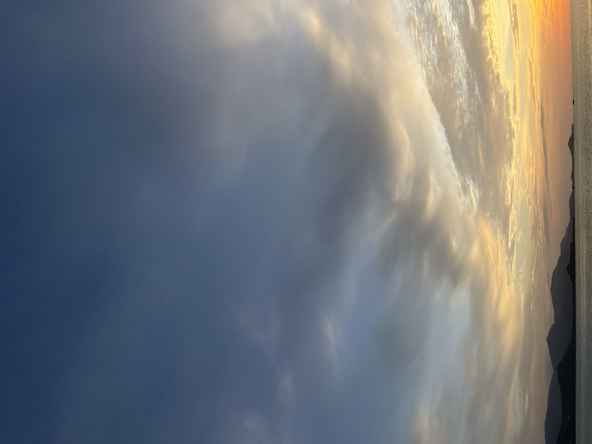 This is a photograph of a dramatic sunset over the ocean, likely taken from a boat or a coastal vantage point in Ko Samui District, Thailand. The scene is dominated by the sky, which displays a gradient of colors from deep blue on the left to fiery oranges and yellows on the right, indicative of sunset.
Large, textured clouds are a prominent feature, catching the warm light of the setting sun and creating a sense of depth and movement. The clouds appear to be illuminated from behind, with bright edges and darker undersides, adding to the dramatic effect.
In the far right of the image, silhouetted mountain ranges are visible against the bright sky, suggesting a coastal or island landscape. The ocean is visible as a calm, gray expanse to the right of the mountains, with a slight ripple on its surface.
The overall mood conveyed by the image is one of awe and tranquility, with the vibrant colors of the sunset evoking a sense of natural beauty and the peacefulness of the evening. The time of day is clearly sunset. There is no visible text in the image.