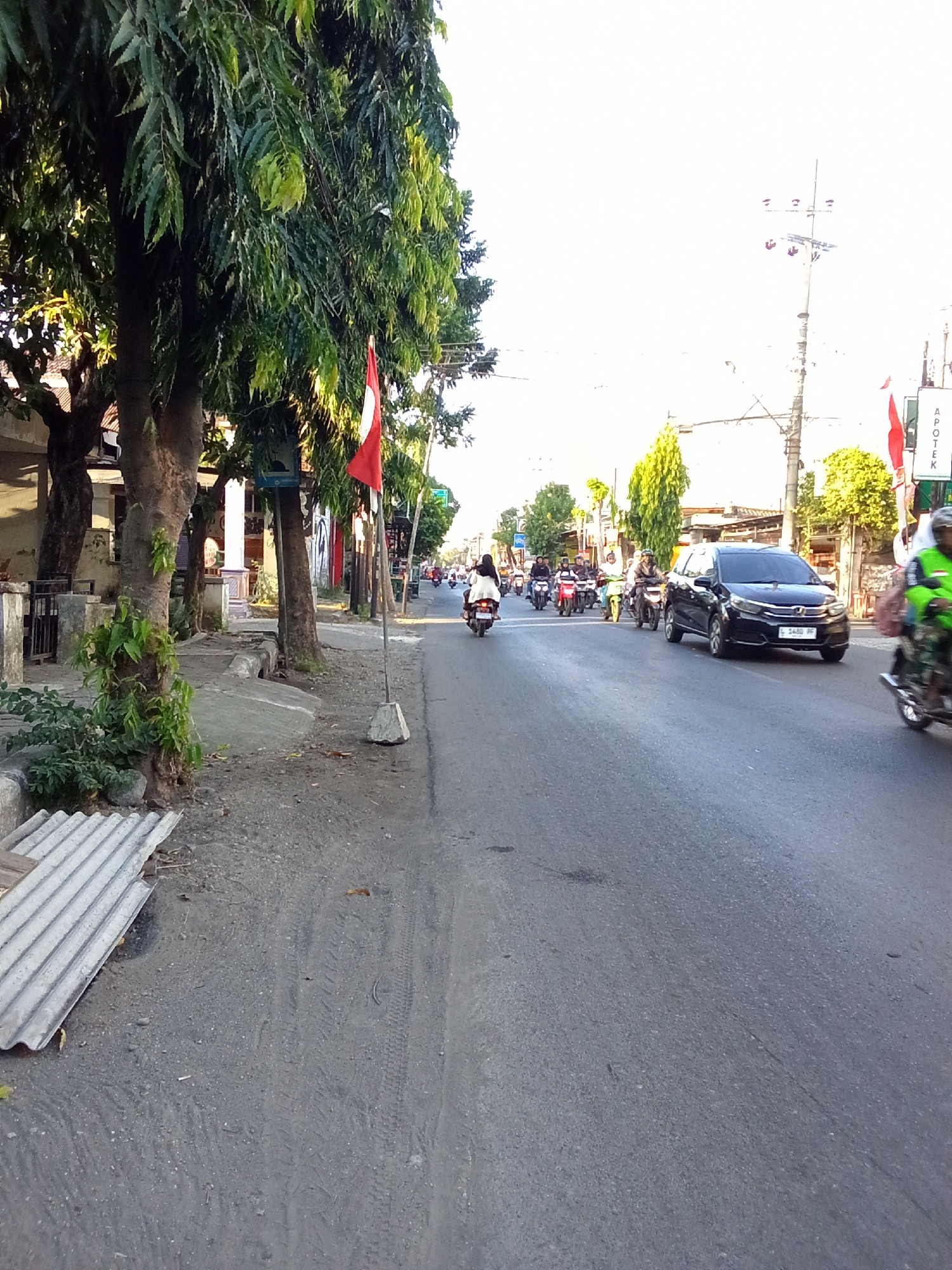 The image depicts a street scene in Kediri City, Indonesia, during the daytime. The perspective is from the side of the road, looking down a paved street. On the left side of the image, there is a sidewalk with trees and some structures. The trees are leafy and provide shade. A red and white Indonesian flag is planted on the sidewalk. The road itself is busy with traffic, primarily motorcycles, and a few cars. In the foreground on the left, there is a pile of corrugated material, possibly roofing sheets. The road surface shows tire marks and some debris. In the background, buildings and power lines are visible, along with more vegetation. The sky is bright and appears to be clear or partly cloudy. There is a sign that reads "APOTEK" which means pharmacy in Indonesian.