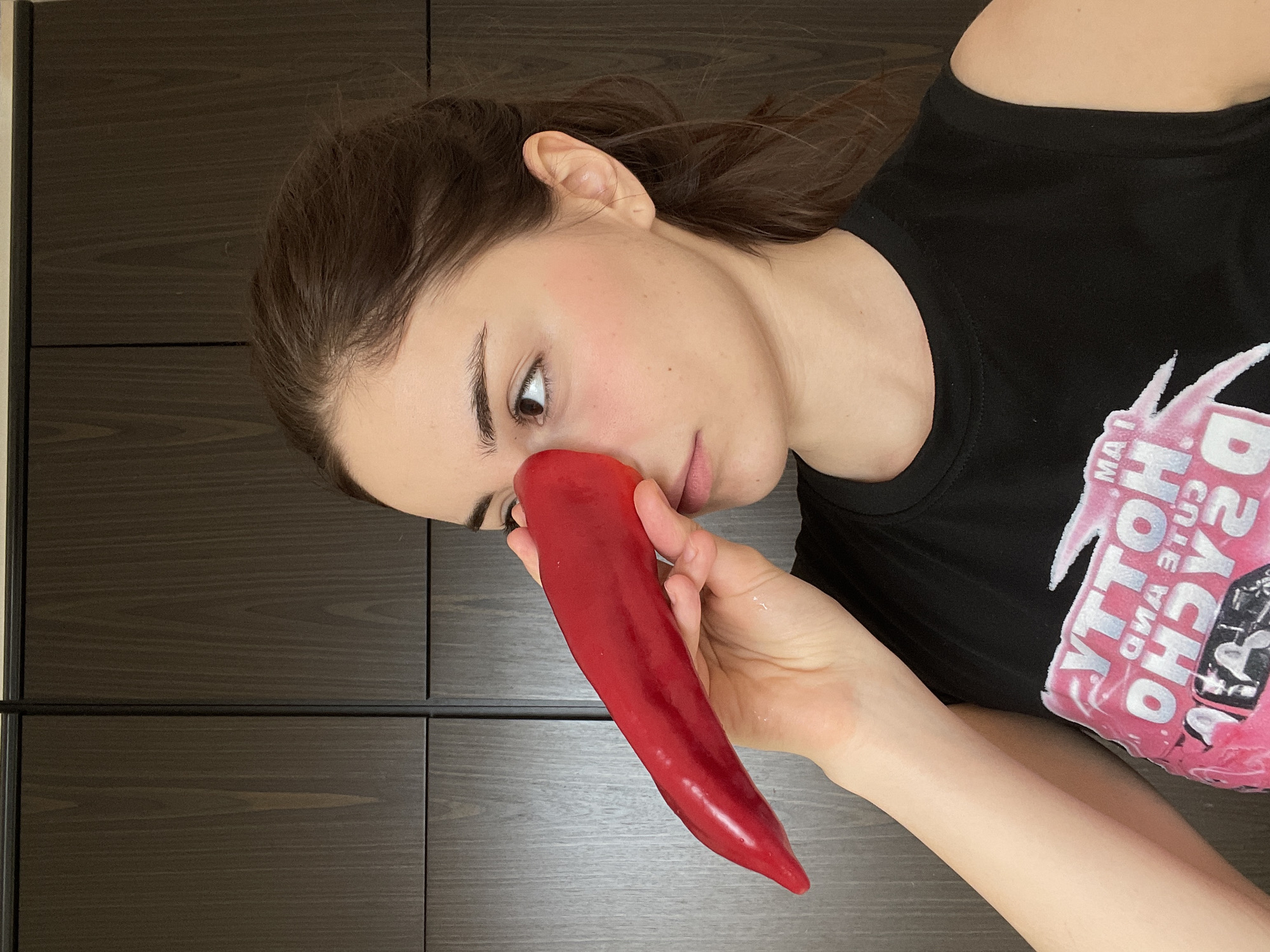 A young woman is depicted from the chest up, positioned diagonally against a dark brown, wood-paneled background, which appears to be cabinetry or wall panels. She has brown hair, visible against her shoulder and partially covering her ear. Her skin has a light blush on her cheek. She is wearing a black t-shirt with pink and white text on it.
In her right hand, she holds a long, vibrant red pepper, bringing it close to her face. The pepper is positioned near her nose and mouth, and she looks intently at it with a focused expression, her right eye visible and gazing towards the pepper. Her lips are slightly parted. The hand holding the pepper is also visible, with wetness or sheen on her fingers and the pepper itself.
The text on her t-shirt is visible in pink and white, appearing reversed. Upon closer inspection and reversal, the text reads: "I AM A CUTE AND HOLY PSYCHO".
The setting appears to be indoors, possibly in a kitchen or living area, suggested by the dark wooden cabinetry. The overall lighting is soft and even. No specific time of day or weather is discernible. The location context is Moscow, Russia.