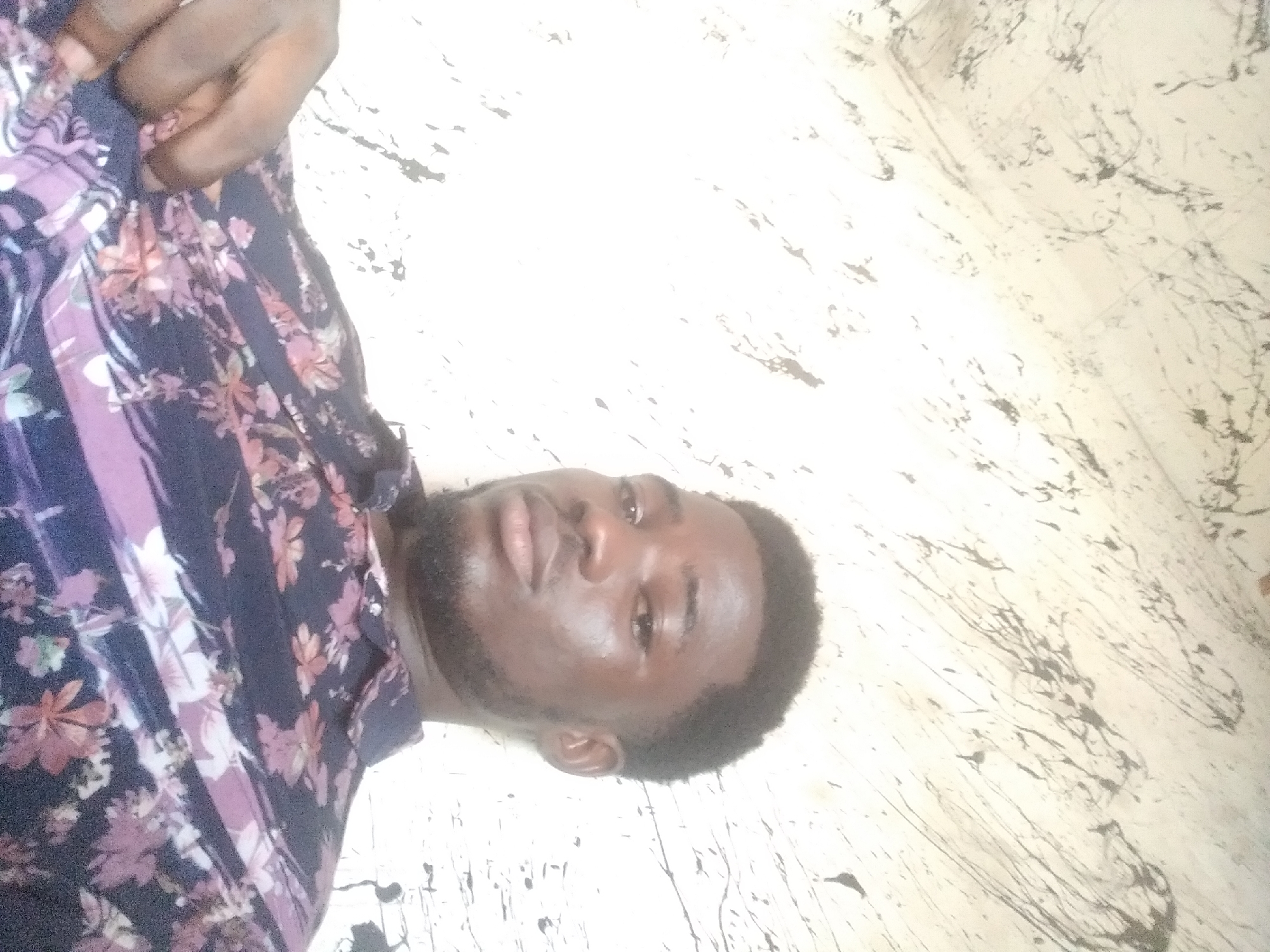 The media file is a close-up, horizontally oriented photograph of a young Black man lying down. He is wearing a dark blue shirt with a floral pattern of pink and purple flowers. His head is turned to his right, and he is looking towards the viewer. He has dark, short hair and a short beard and mustache. His eyes are open and he appears relaxed. The background is a light-colored surface with a marbled or patterned texture, possibly a floor or wall, with dark, irregular markings. The lighting suggests it is daytime, possibly indoors given the bright, even illumination and lack of shadows. There is no visible text in the image. The overall mood is casual and intimate, perhaps a selfie taken in a personal space.