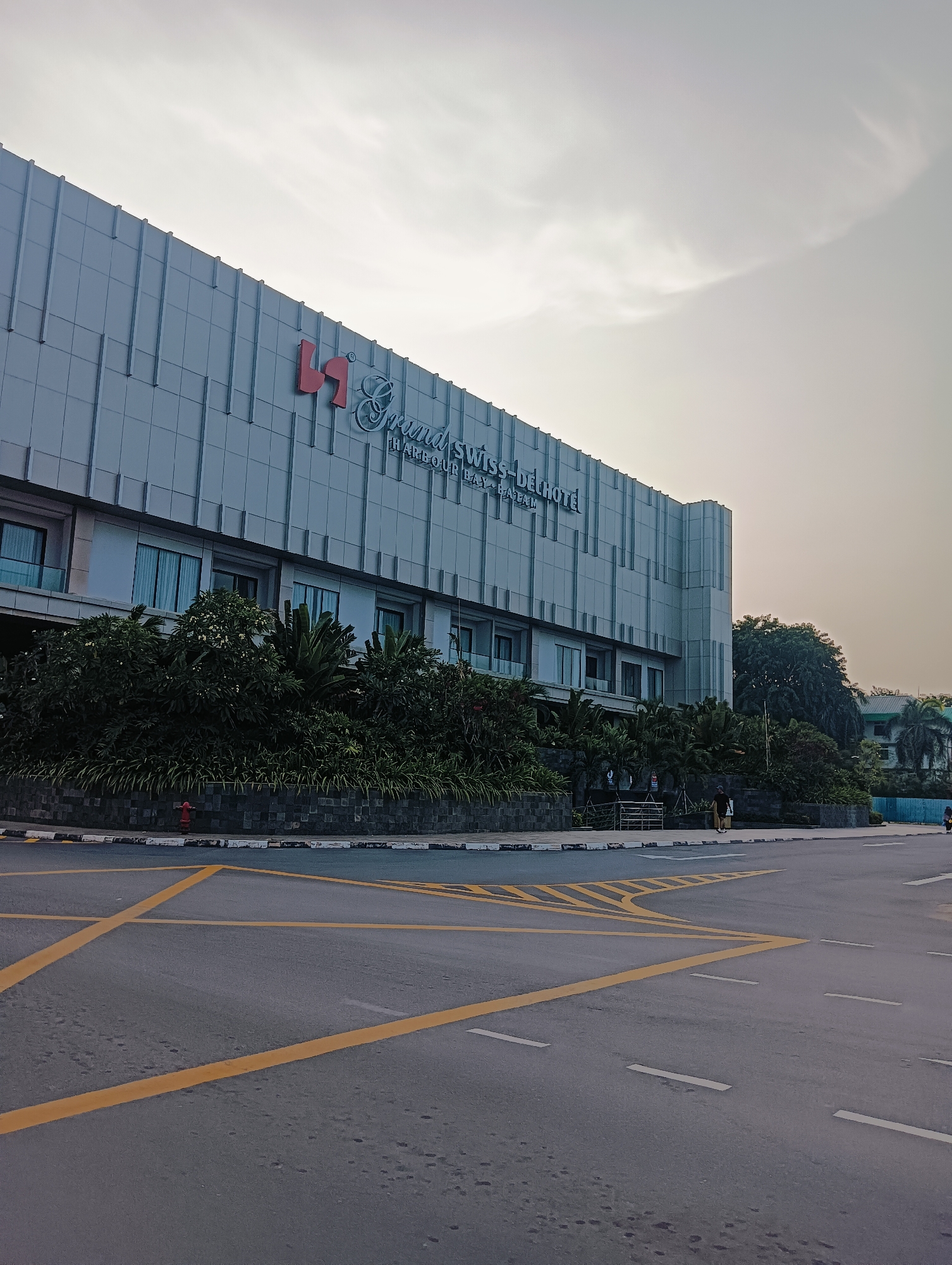 The image shows the exterior of the Grand Swiss-Belhotel Harbour Bay in Batam. The hotel building is modern with a light-colored facade. In the foreground, there is an asphalt road with yellow lane markings. Lush green vegetation, including trees and bushes, is planted along the front of the hotel. A red fire hydrant is visible near the sidewalk. A few people are walking on the sidewalk in the distance. The sky is overcast with some clouds.