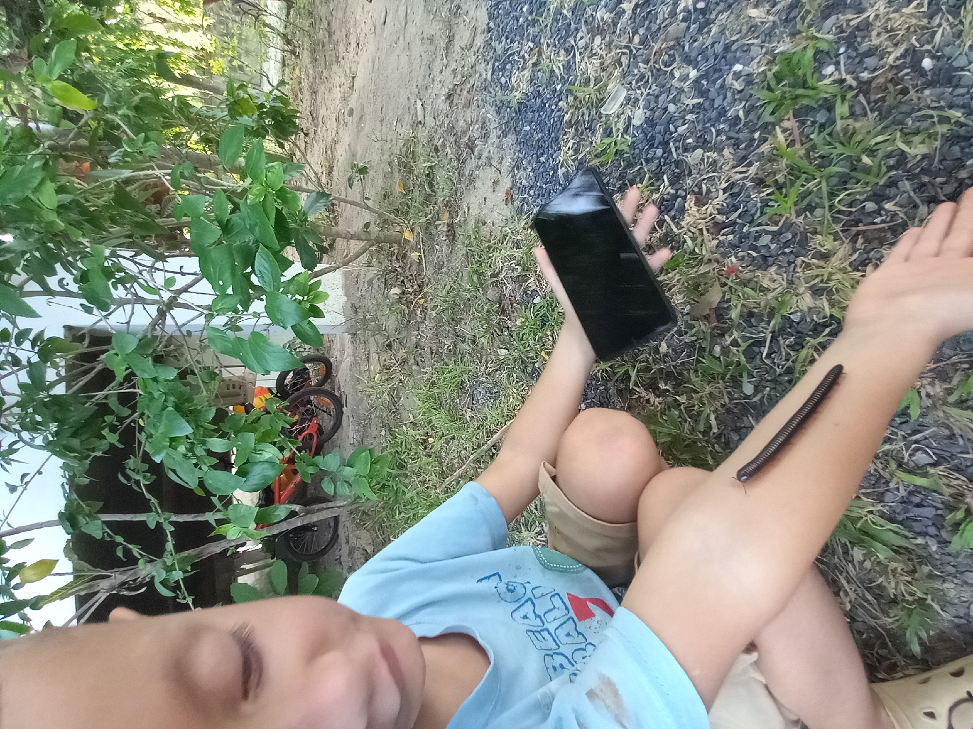 In Baan Mae Nam, Thailand, a close-up, outdoor, eye-level shot shows a young child sitting on the ground. The child, wearing a light blue t-shirt with graphics and tan shorts, is holding a black smartphone in their right hand. A large millipede is crawling on the child's left arm. The background features green foliage and two bicycles parked behind a tree. The ground is covered in gravel and patches of grass.