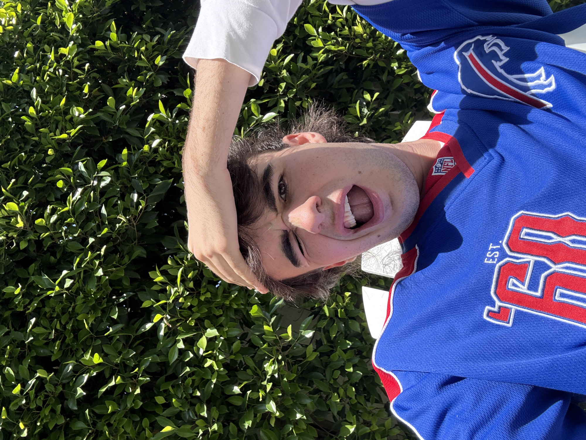 A young man in a blue jersey with red and white trim is held upside down in front of a green hedge. The man's open mouth, teeth, and tongue are visible. The jersey features the logo of the Buffalo Bills NFL team. A second person's hand, wrist, and sleeve are visible, holding the first man upside down. The location is likely an outdoor space with foliage, possibly in Los Angeles, based on the prompt, but there are no specific visual cues in the image to confirm the location.