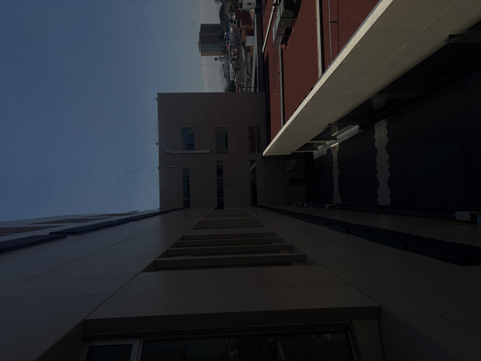 The image shows a low-angle view of two buildings in Puebla, Mexico. The building on the left is taller and has a pattern of windows and horizontal architectural details. The building to the right has a red roof and a white overhang. In the distance, other buildings are visible against a blue sky.