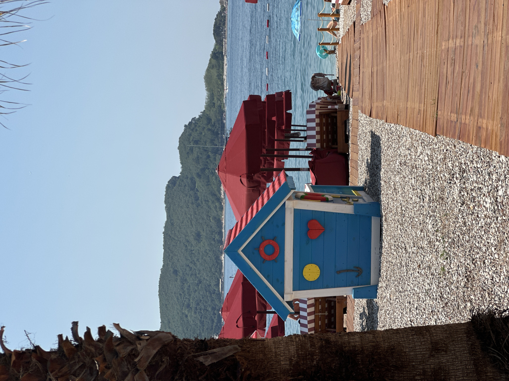 This is a vertical, eye-level shot of a beach scene in Budva, Montenegro. The foreground is dominated by the rough texture of a palm tree trunk. In the mid-ground, a small, brightly colored wooden playhouse, painted blue with red and white striped accents, is the focal point. It is decorated with a red life preserver, a red heart, and a yellow circle, and has a small brown anchor-shaped hook. Behind the playhouse, several closed red beach umbrellas are arranged in rows.
Further back, a wooden boardwalk runs parallel to the shoreline, with a scattering of small, white pebbles on either side of it. The sea, a vibrant blue, stretches out to the horizon. A distant, tree-covered mountain range forms the backdrop. The water is calm, with a few buoys indicating a swimming area. A couple of beach umbrellas are open further down the beach, and a few people are visible relaxing on chairs. The sky is clear and blue, suggesting a sunny day. The overall atmosphere is one of a peaceful, summer day at the beach. No text is visible in the image.