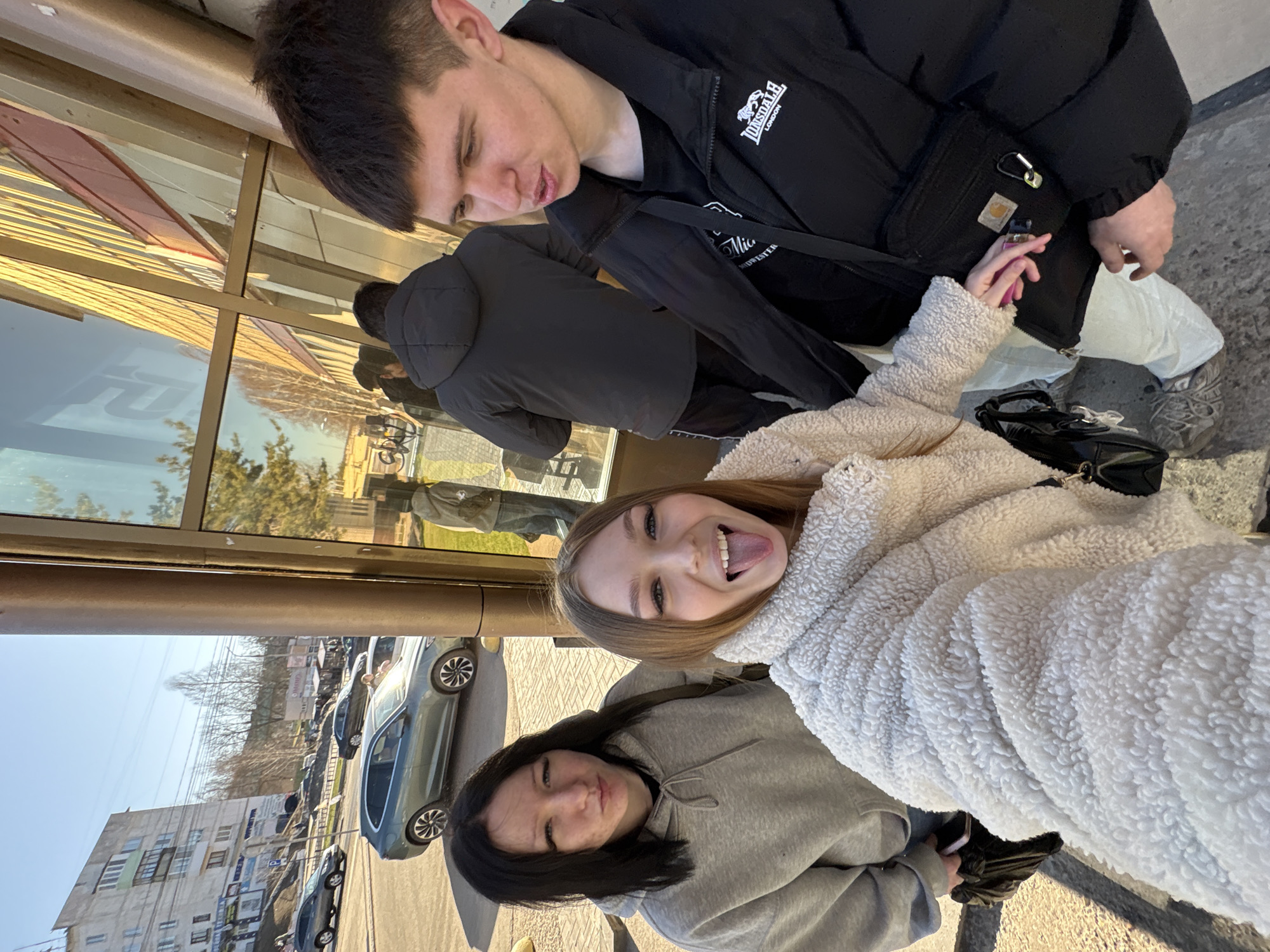 The image depicts a group of four individuals in an outdoor setting, likely in the city of Chaikovsky, Russia, based on the urban backdrop and architectural style. Three are positioned closer to the camera and are in focus.
A young woman with light brown hair is in the foreground, wearing a white, fluffy jacket. She is sticking her tongue out. Next to her is a person wearing a gray hoodie and with dark hair. Above them is a young man wearing a black jacket with a Carhartt pouch attached to it.
The fourth person, wearing a dark jacket with a hood, is further back.
The background shows a street scene with buildings and parked cars. The reflection of trees and buildings can be seen in the windows to their side.