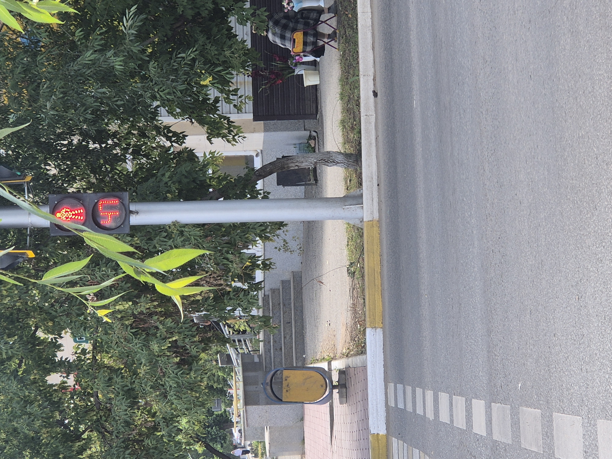 A traffic light with a red pedestrian signal depicting a walking figure and a countdown timer showing "40" is mounted on a pole. The traffic light is positioned near a tree with green leaves. In the background, there are building facades, stairs, a trash can, and a person sitting on a chair. The foreground shows a pedestrian crossing with white square markings on the asphalt and a yellow and white curb. The scene appears to be a street in the city of Kokshetau, Kazakhstan.
