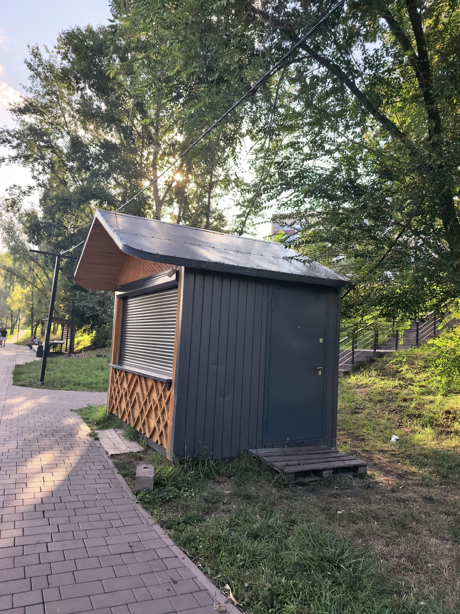 This is a photograph of a small, dark-gray kiosk with a sloped, dark-gray metal roof, accented with light-brown wooden trim under the roof overhang. The kiosk has a closed roll-up door covering the serving window, which is framed by a decorative diamond-patterned wood lattice. To the right of the serving window, there is a dark-gray door with two lock mechanisms. A wooden pallet serves as a makeshift step in front of the door. The kiosk is situated on a grassy embankment next to a paved walkway made of rectangular gray bricks. The walkway runs diagonally from the lower-left to the mid-left of the frame. Trees with green foliage are abundant in the background, with sunlight filtering through the leaves. In the distance, a set of stairs with metal railings ascends a grassy slope to the right of the kiosk. The scene appears to be a public park or recreational area.