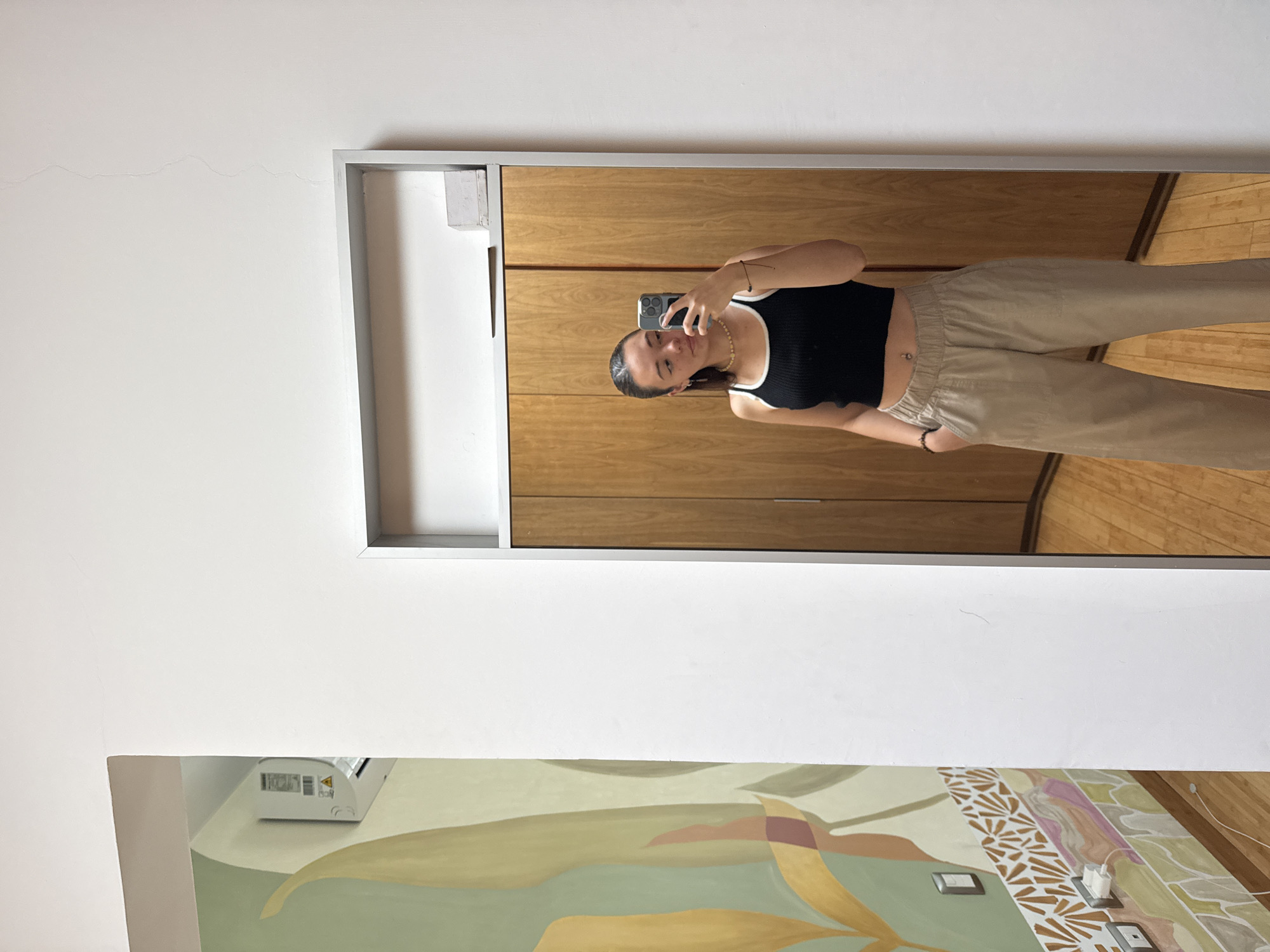 A young woman stands in front of a mirror, taking a selfie with her smartphone. She wears a black tank top with white trim and light beige pants. Her hair is pulled back, and she is wearing a bracelet and necklace. Behind her in the reflection are wooden cabinets and flooring. Below the mirror and slightly to the left is a painted mural of abstract floral shapes. The wall above and around the mirror is white. The location is identified as San Bernardino Tlaxcalancingo, Mexico.