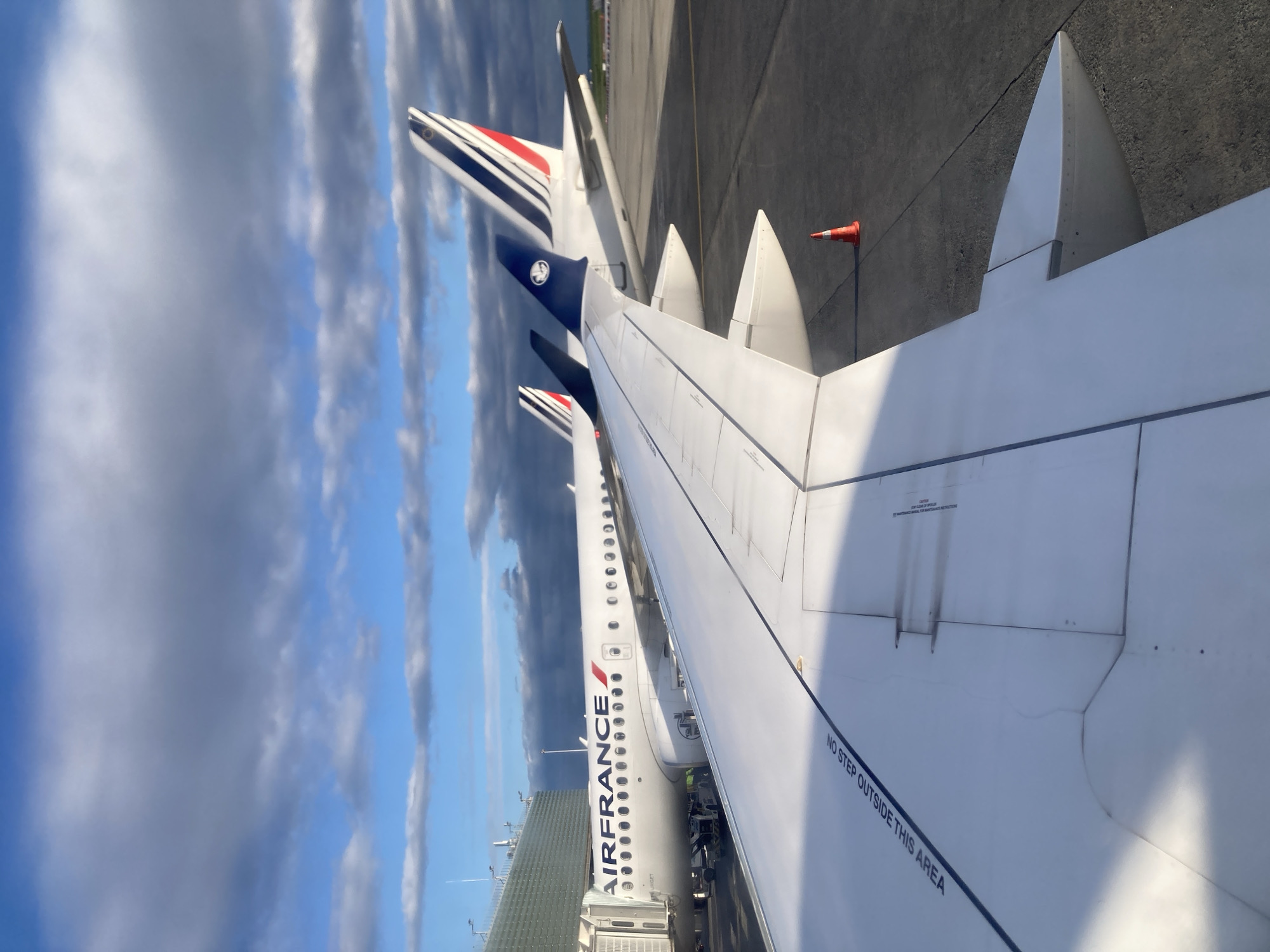 The photograph shows a view from an airplane wing, likely taken at Charles de Gaulle Airport (CDG) in Le Mesnil-Amelot, France. Two Air France planes are visible, their fuselages bearing the airline's name and logo. The tail fins are painted with the French tricolor. The focus is on the wing of the aircraft from which the picture was taken, displaying a "No Step Outside This Area" marking. The sky is filled with clouds, and a section of airport tarmac can be seen alongside a small orange cone. The buildings surrounding the airport are visible in the background.