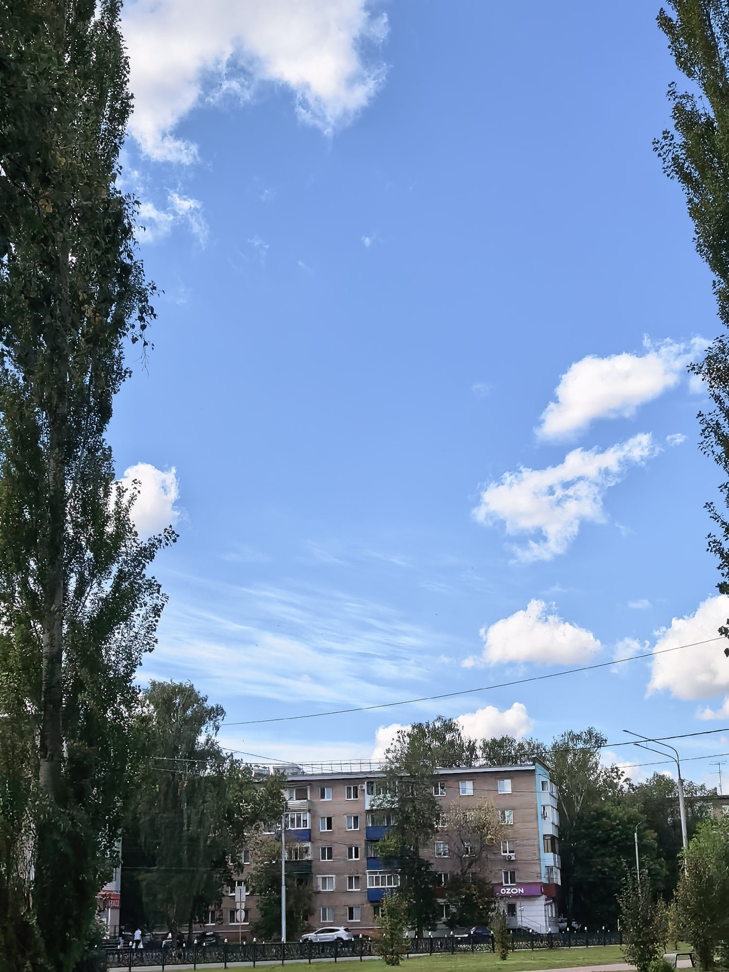 The image shows a view of a residential building in Sterlitamak, Russia, framed by tall trees on either side. The building is a multi-story apartment block with a light brown and blue facade. There are several windows visible, some with air conditioning units. A sign for "OZON" is visible on the ground floor of the building. The sky is blue with scattered white clouds and some wispy cirrus clouds. In the foreground, there is a street with cars and people walking, separated by a fence and a grassy area with young trees. The scene appears to be during daytime, under a clear sky.