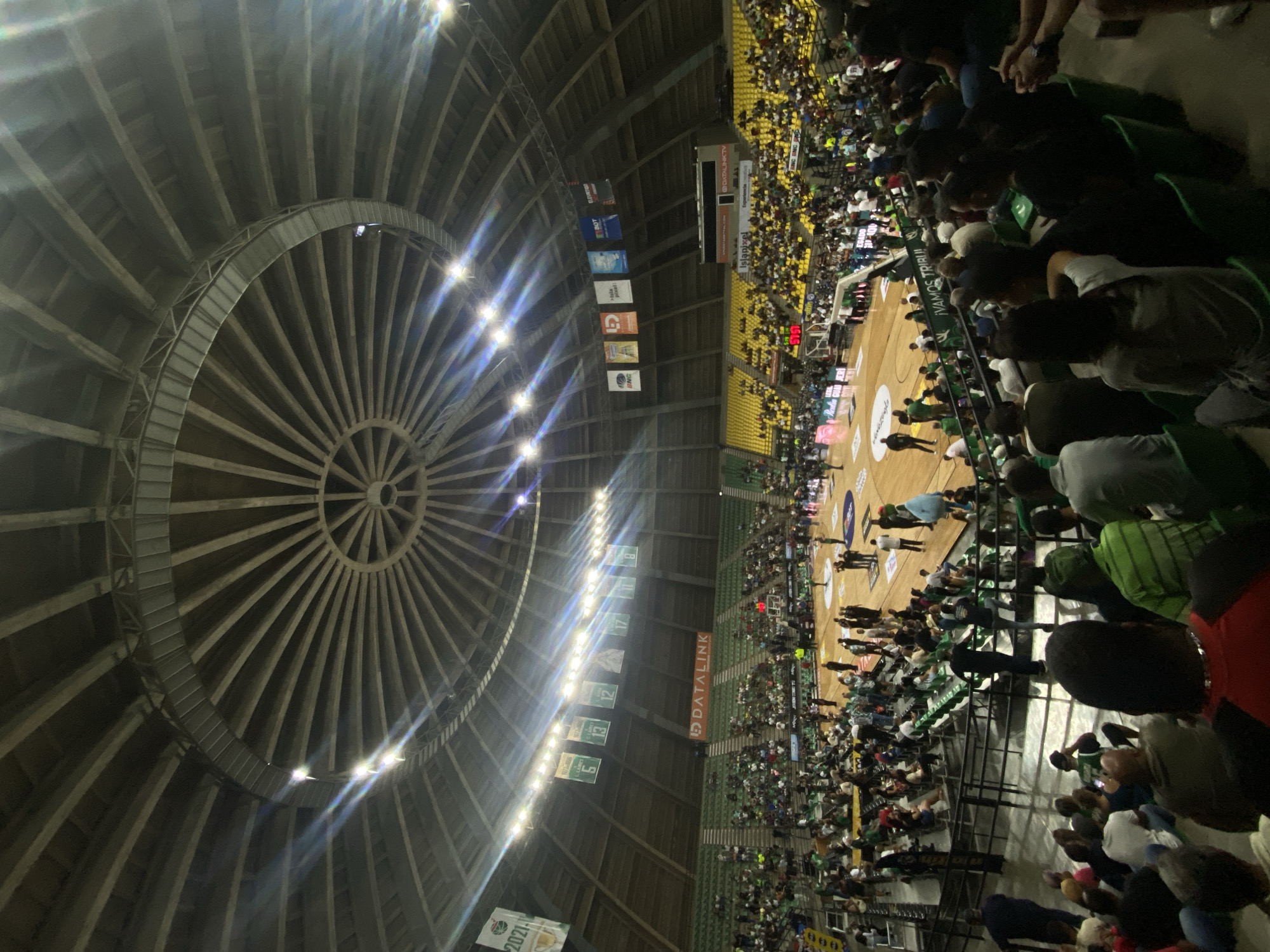 The image shows the Polideportivo Ciudad de La Asunción, a basketball arena in La Asunción, Venezuela, during a game. The arena is packed with spectators. The distinctive circular ribbed roof structure is visible. Banners hang from the ceiling. Players are on the court. Fans are seated in the stands.
