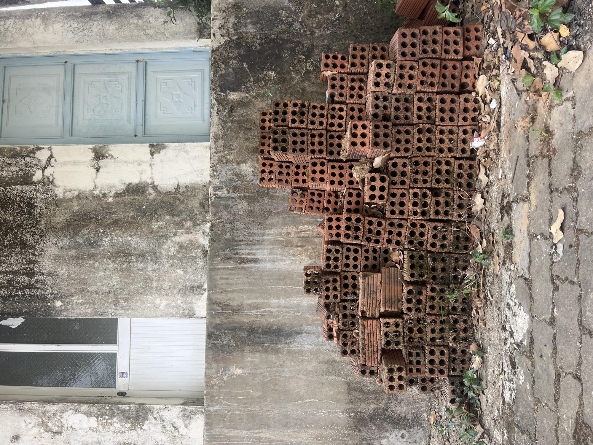 The image presents a vertical view of a building exterior and adjacent walkway, likely in Bờ Y, Vietnam. A stack of red clay bricks occupies a significant portion of the right side, partially overlapping the walkway. The bricks are riddled with cylindrical holes and appear weathered.
To the left, the building facade features a combination of textures and materials. There is a light blue, ornamental window near the top and a white security window near the bottom. The wall surface is a mottled gray, with variations in shade suggesting moisture or age. The walkway is concrete. There are leaves and small plants near the bricks.