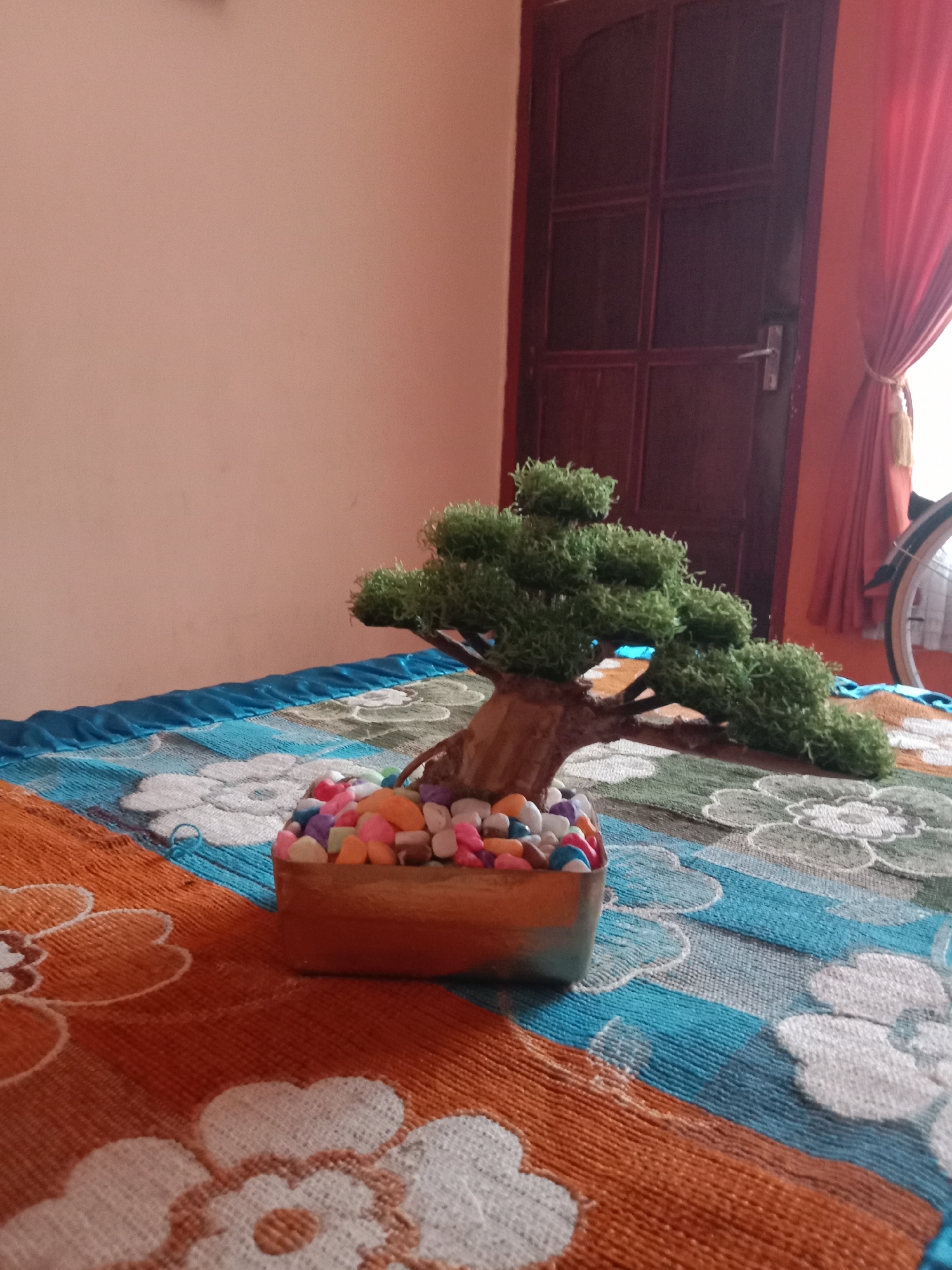 This is a close-up, eye-level, indoor photograph taken on a bed with a patterned blanket. The blanket features large white floral designs on a background of orange, blue, and green stripes. In the foreground, a small, artificial bonsai tree is placed in a rectangular pot filled with colorful, small stones. The tree has a brown, textured trunk and bushy green foliage. Behind the bed, a dark brown door with glass panes is visible, along with a reddish-orange curtain and a portion of a bicycle wheel with spokes. The wall behind the bed is painted a light, peachy color. The scene appears to be in a bedroom setting.