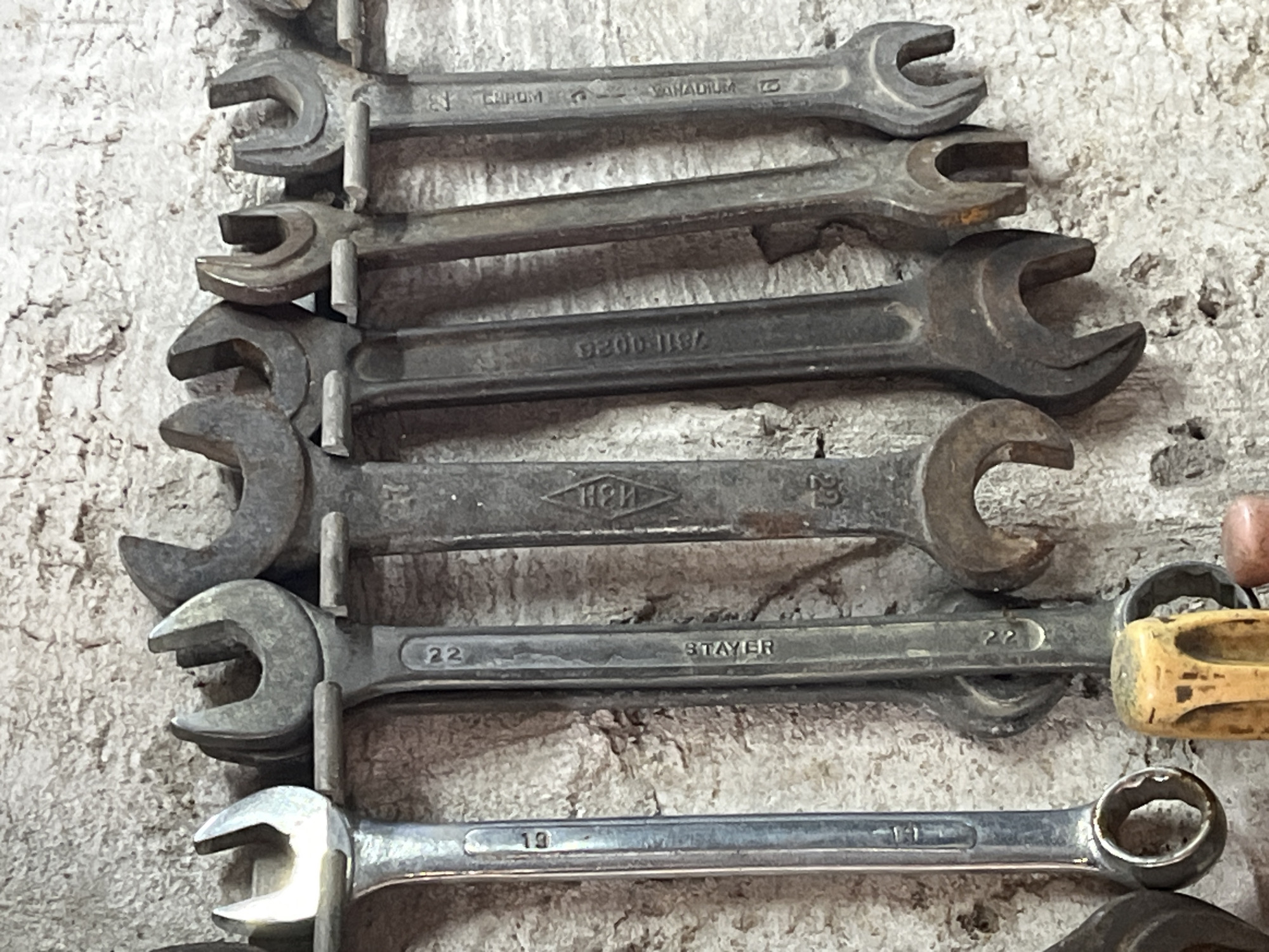 An overhead view of a wall-mounted rack holding several wrenches of varying sizes and conditions. The wrenches are arranged horizontally, with their handles pointing downwards. The wall appears to be made of concrete or plaster, with a rough, textured surface. The lighting suggests an indoor environment, possibly a workshop or garage. The specific location of Krasnyye Barrikady, Russia is not determinable from the visual information present in the image.