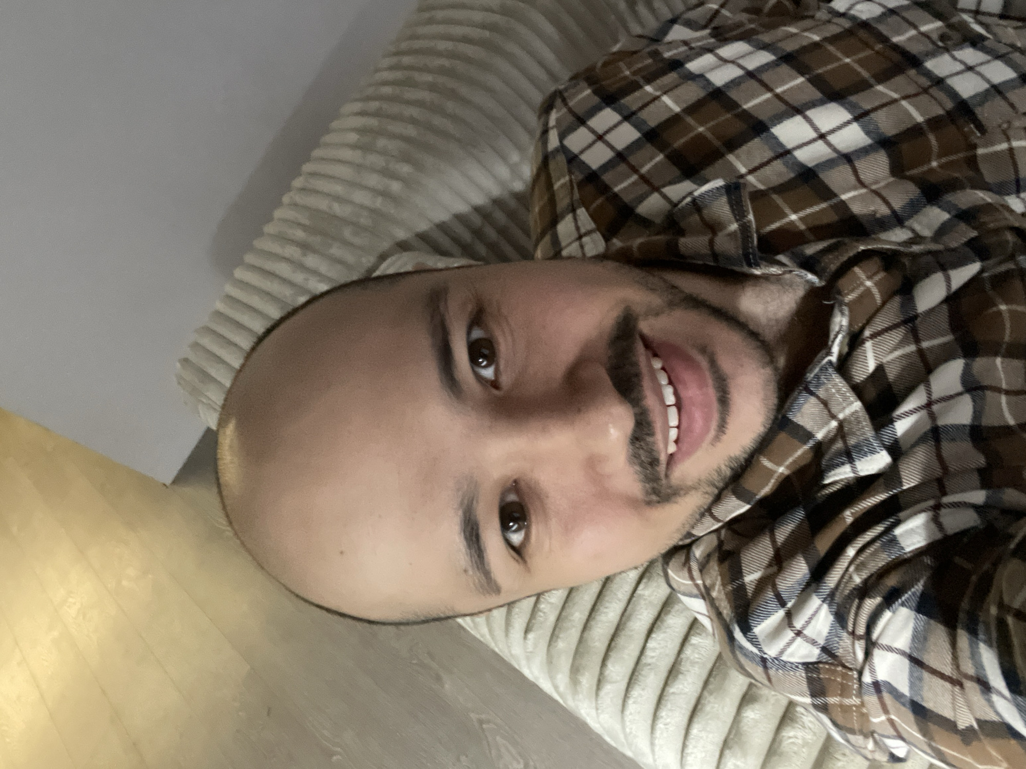 A man with a shaved head and a mustache is lying on a light-colored, ribbed sofa, wearing a brown and white plaid shirt. The background shows a glimpse of light-colored flooring, possibly wooden. The image does not provide information about the location in Izhevsk, Russia.