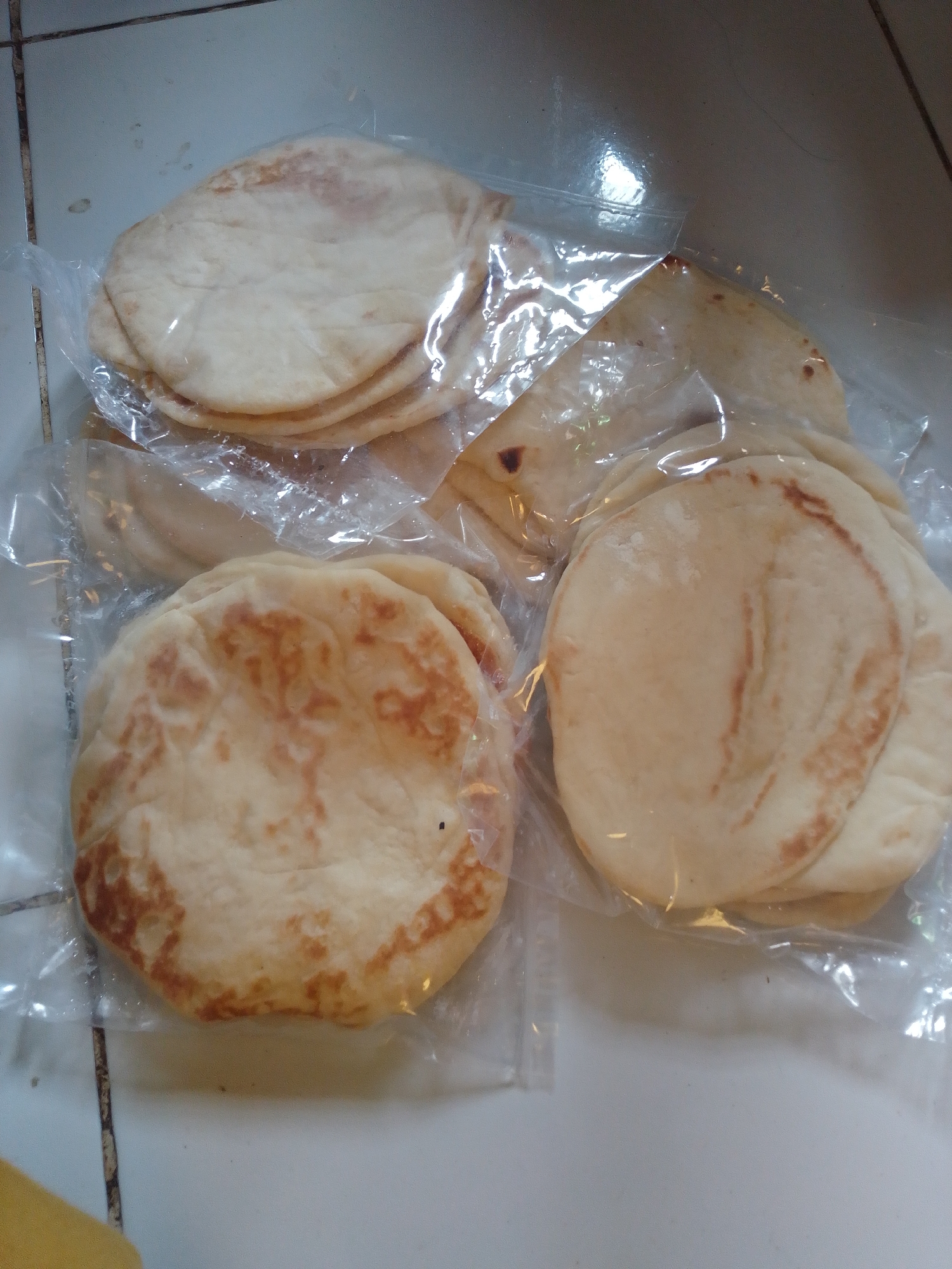 A top-down, medium shot shows a stack of pita breads inside a clear plastic bag, resting on a tiled surface. The pita breads are round and flat, with some areas of light browning and a slightly puffed texture. The plastic packaging is slightly creased and reflective, obscuring some of the detail. The tiled surface appears to be light-colored, possibly ceramic, with visible grout lines running horizontally and vertically. The background is dimly lit, with a hint of what might be a wall or another surface. The scene is static, with no people or animals present. The lighting is soft, with a slight glare on the plastic packaging. The overall impression is of a domestic setting, likely a kitchen counter or pantry, in Panyingkiran, Indonesia.