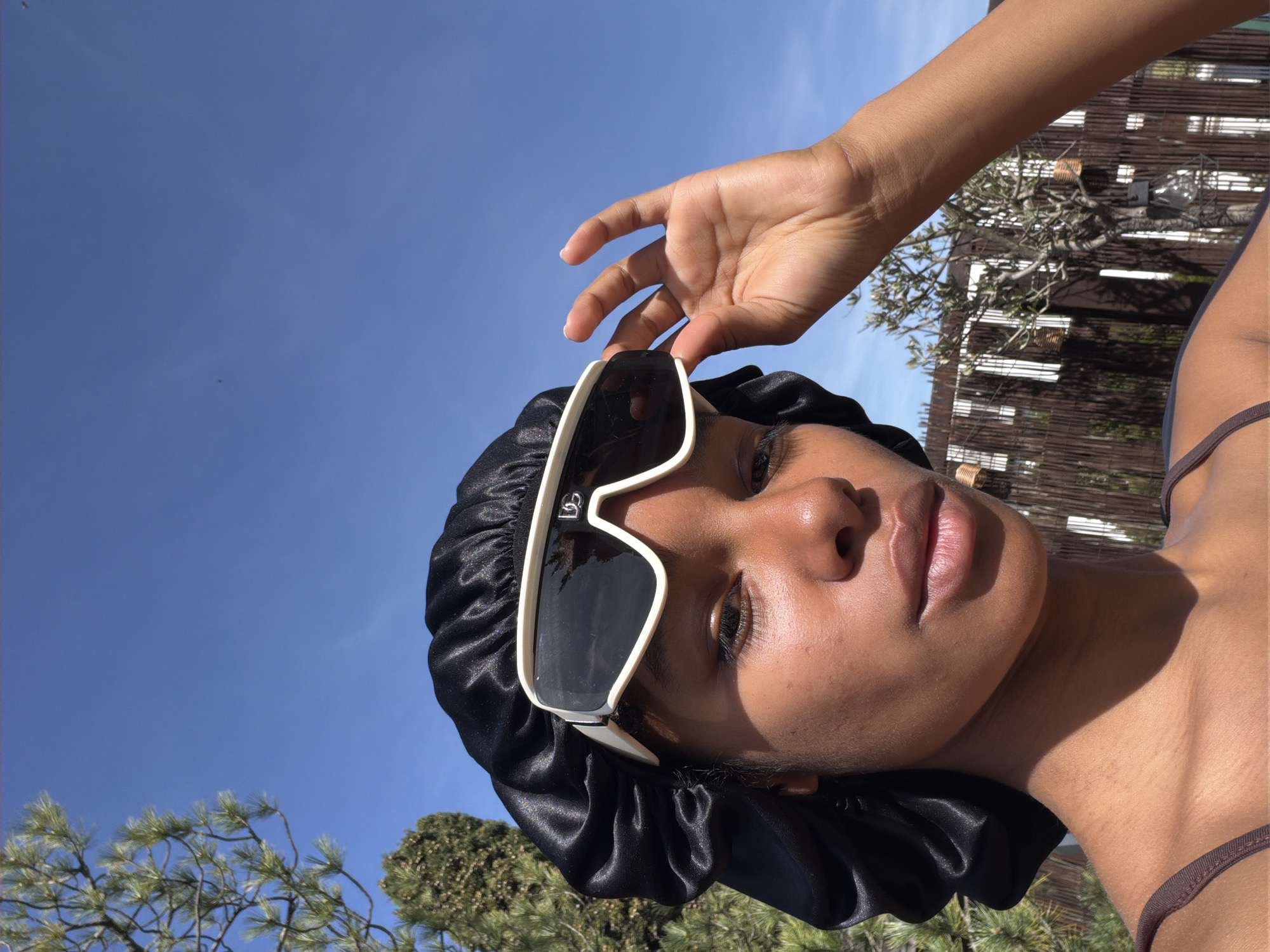 A medium close-up shot depicts a woman wearing a black silky bonnet and large white-framed sunglasses. Her right hand is raised, with fingers lightly touching the sunglasses' frame. The woman has brown skin, full lips, and her gaze is directed slightly upwards.
Behind her is a clear blue sky. Green trees and shrubs are visible in the lower left corner. A light-colored fence or wall is visible to the right, with a bare tree or bush behind it. The environment suggests an outdoor setting. The location is unknown.