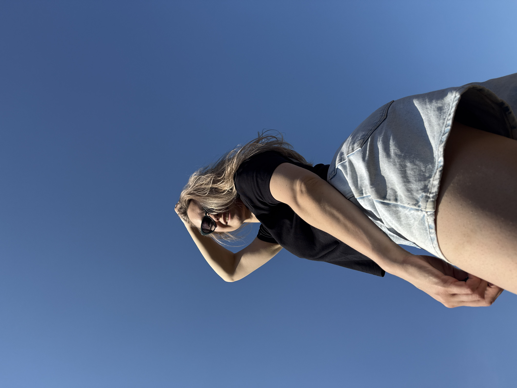 A low-angle shot of a woman wearing a black t-shirt and light-wash denim shorts, framed against a clear, bright blue sky. She wears sunglasses and has blonde hair. Her right arm is raised, as if shielding her eyes from the sun. The location is not discernible from the image, so it's impossible to confirm if it is in Chaikovsky, Russia.