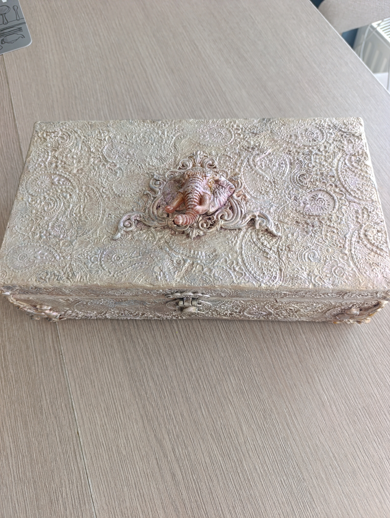 A vintage, ornate, rectangular wooden box with a textured, decorative surface in a pale gold and cream color scheme sits on a light brown wooden table. The box has a hinged lid with a metal clasp in the center. The lid is adorned with a raised, decorative design featuring an elephant-like motif in a pinkish-brown color, surrounded by an intricate, embossed pattern of swirling paisley and floral elements. The box appears to be a decorative storage container. The environment suggests an indoor setting, possibly a home. There is no identifiable location in the city of Toruń, Poland, or any people or actions present in the image.