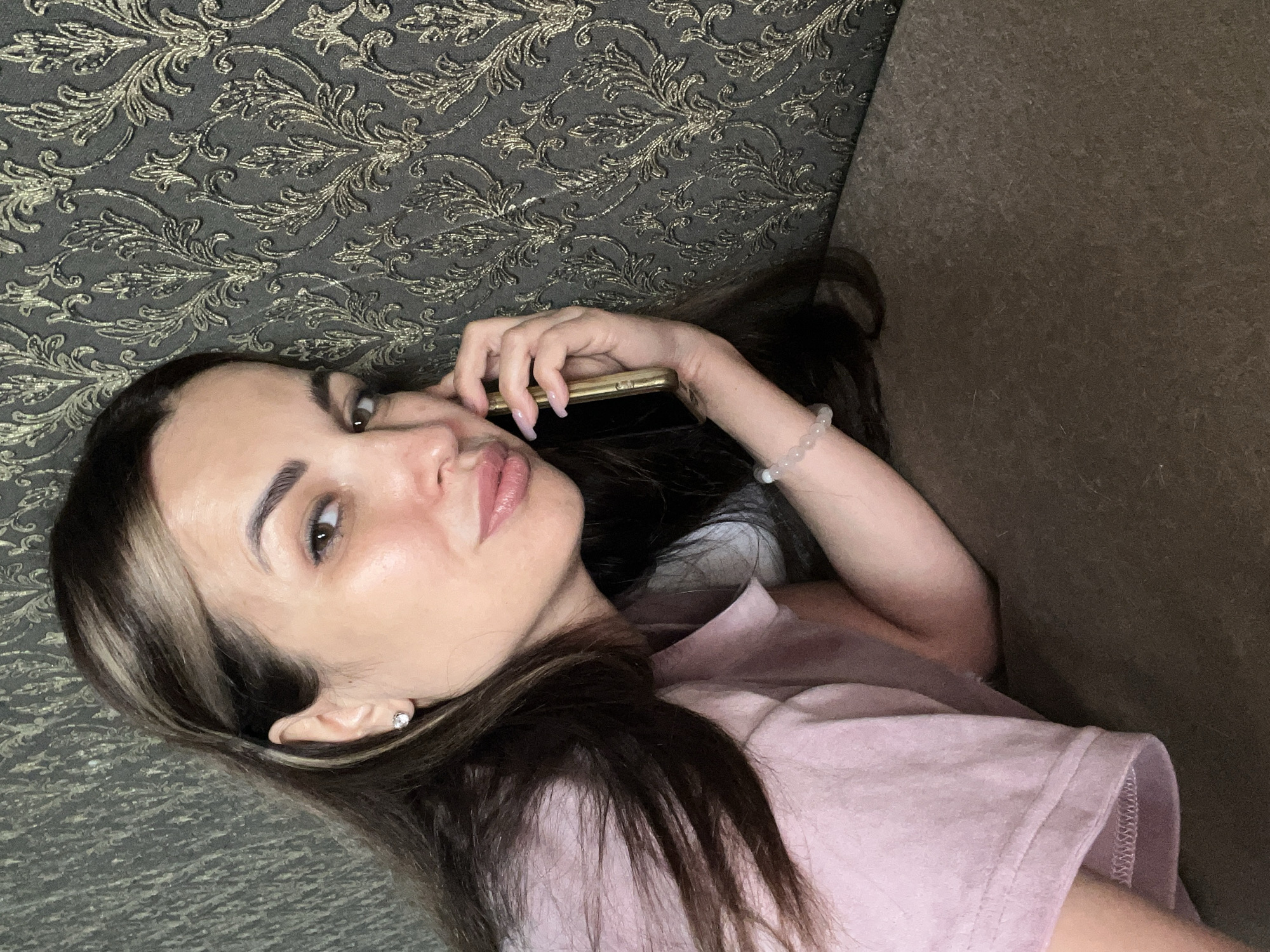 A woman with long, dark hair with blonde highlights is lying down, leaning against a wall with patterned wallpaper. She is wearing a pink t-shirt and a bracelet on her wrist. She is holding a golden phone to her ear, appearing to be in a conversation. Her gaze is directed towards the camera. The background consists of the textured wallpaper and a dark-colored surface that she is resting on.