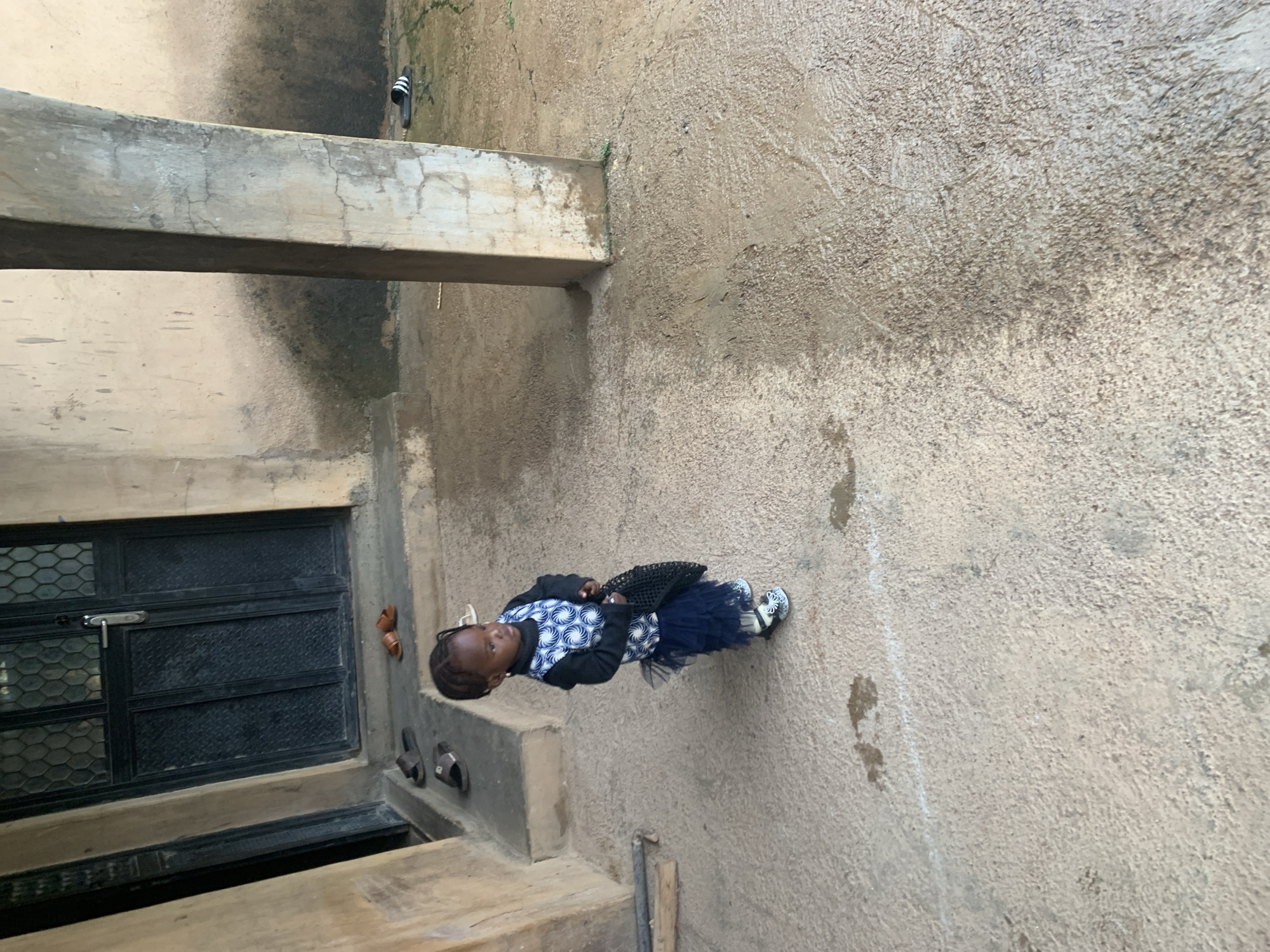 The image shows a young child standing on concrete steps outdoors. The child is wearing a dark blue tutu skirt and a white and blue patterned top with black sleeves, along with white shoes. The child is holding a black handbag. To the left of the child is a dark metal grate covering an opening. Above and to the left of the child, a concrete beam runs horizontally. The wall behind the child is rough and textured concrete. Several pairs of sandals are visible on the steps near the child. The scene suggests a casual, everyday setting. The lighting indicates it is daytime.