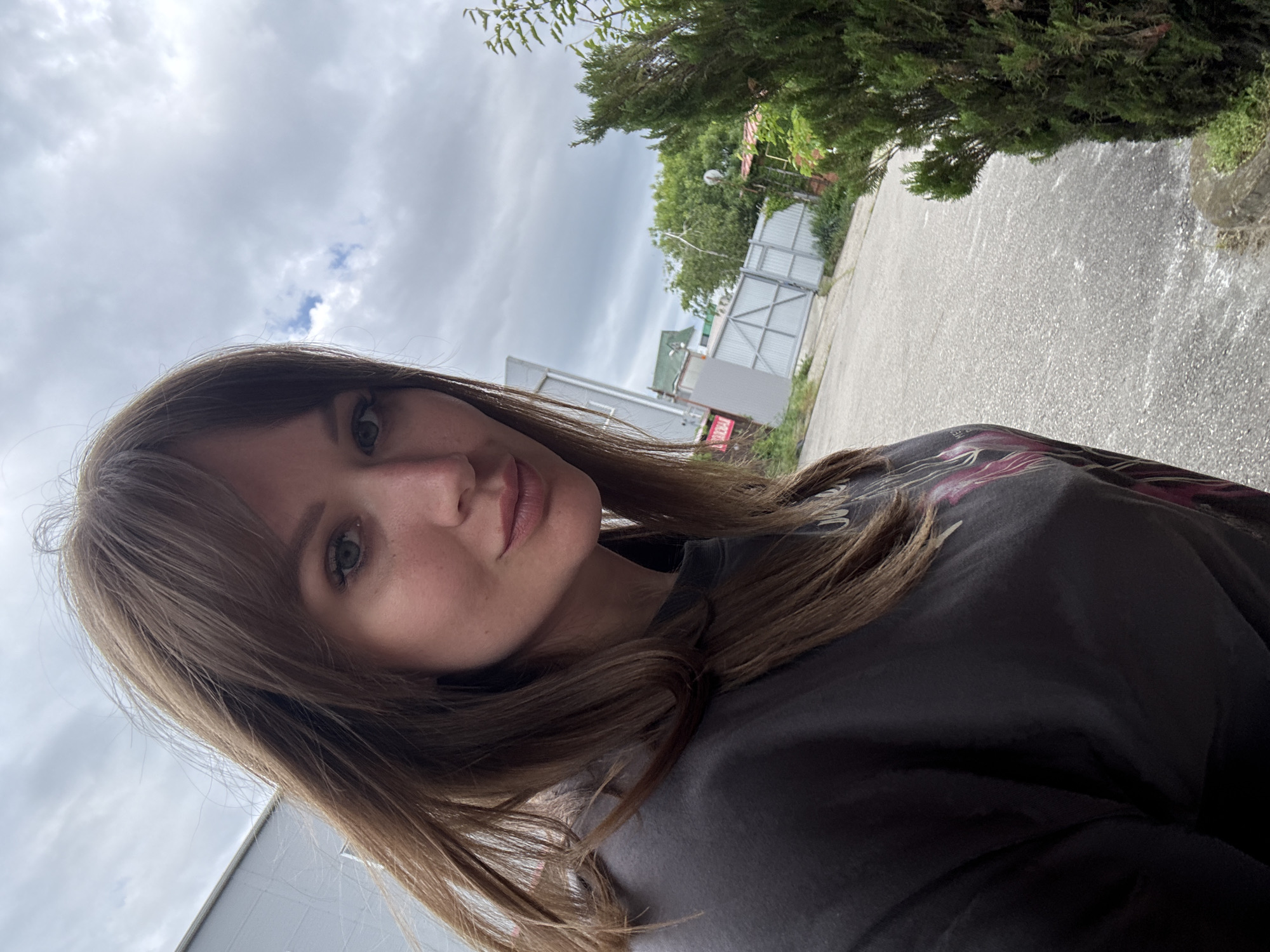 A photograph shows a woman with brown hair and green eyes, positioned in the foreground with a slight tilt. She wears a black shirt. Behind her is an outdoor scene in what appears to be the city of Khadyzhensk, Russia, indicated by signs in Russian. A grey road extends into the background, with a white fence and buildings visible along the side. Trees and greenery are present. The sky is overcast.