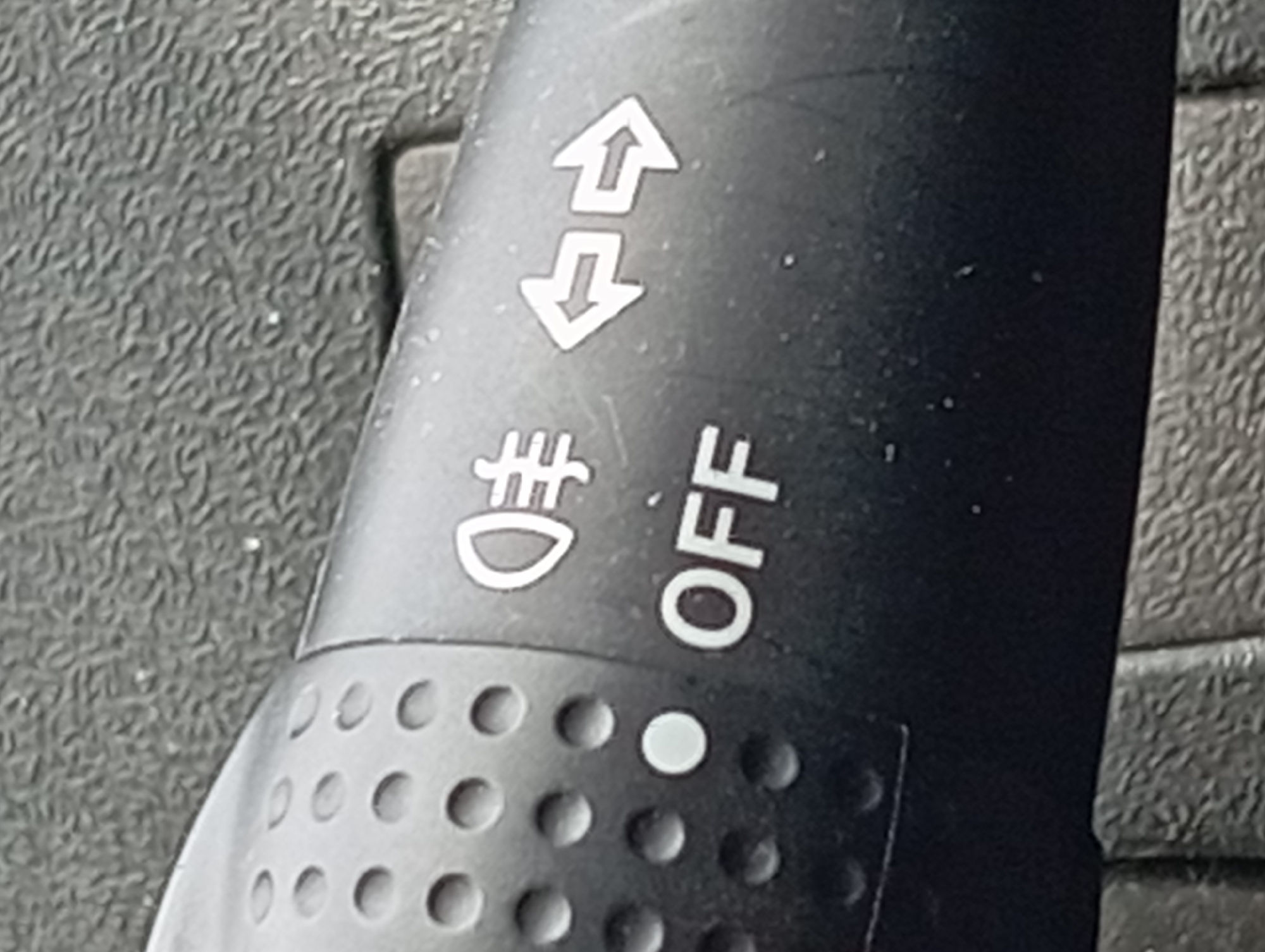 The image is a close-up, slightly angled, eye-level shot of a black car's multi-function turn signal stalk. The stalk has embossed circular dimples on its left side and white symbols and text on its right. A prominent symbol depicts a headlight with vertical lines to its right, indicating the headlight function. Below this symbol is the word "OFF" in capital letters. To the right of the headlight symbol are two opposing arrows, one pointing left and the other pointing right, representing the turn signal indicators. The background is a textured, dark gray surface, likely part of the car's interior. The lighting is somewhat harsh, casting subtle shadows. There are no people or animals visible in the image, and no specific landmarks of Nitra, Slovakia, can be identified. The focus is entirely on the details of the car's stalk.