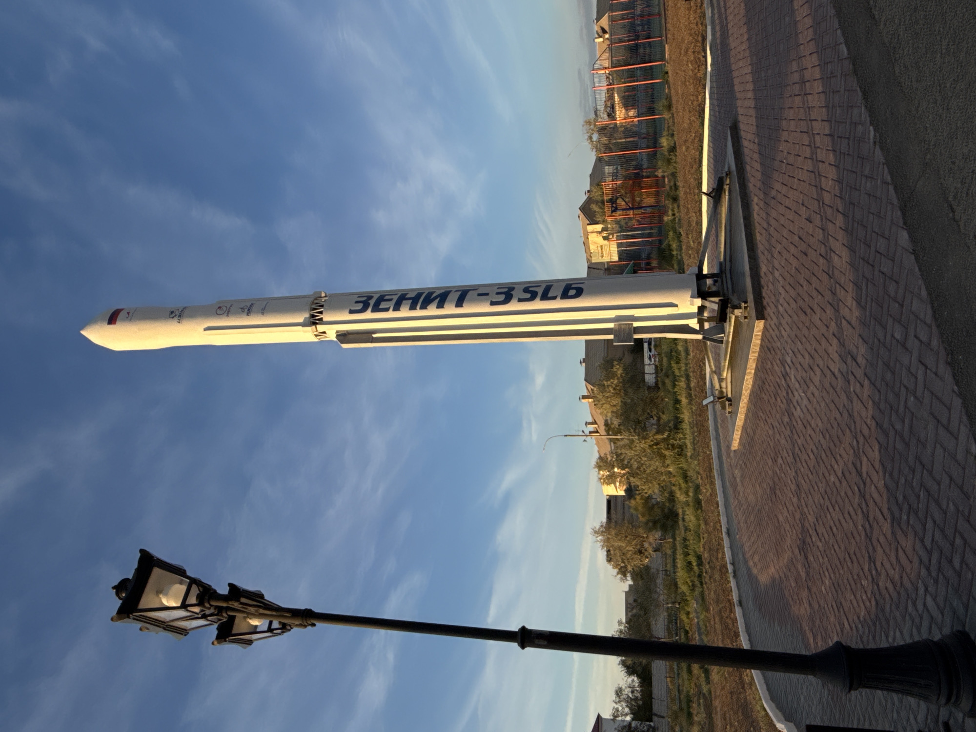 A monument of the Zenit-3SLB rocket stands vertically on a pedestal in an outdoor paved area in Baikonur, Kazakhstan. The monument is white with blue lettering on its side that reads "ЗЕНИТ-3SLB". To the left of the rocket is a black double lamppost. The background shows low-rise buildings, trees, and a sports field with red fencing. The sky is blue with scattered clouds. The sun is low, casting long shadows on the ground. The scene appears to be a public space or park. There are no people visible in the image.