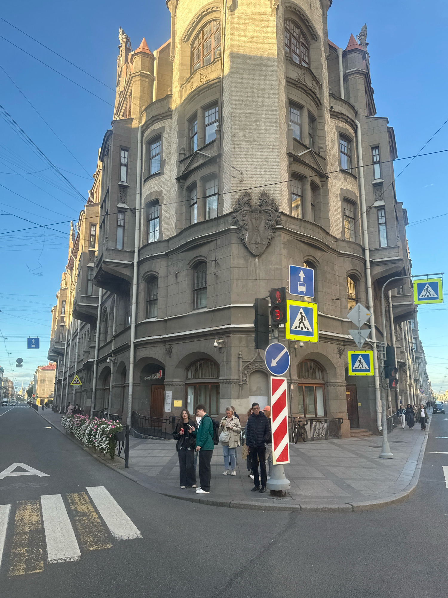 The image depicts a street corner in Saint Petersburg, Russia, with a focus on an ornate, multi-story building characterized by its detailed architectural features, including arched windows, balconies, and decorative elements. The building appears to be of historical significance, possibly constructed in the Art Nouveau or a similar architectural style prevalent in the city. The sky is clear and blue, indicating fair weather, and the sunlight casts shadows, suggesting it is either morning or late afternoon.
On the sidewalk in front of the building, several pedestrians are visible. A group of four people are standing near a traffic light and a series of road signs. One person is wearing a green jacket, another a black jacket, and two are wearing darker clothing. Further down the street, more people are walking and some are standing near a row of planters filled with flowers.
The street itself features a pedestrian crossing marked with white and yellow stripes. Traffic lights are present, along with various road signs, including directional signs and pedestrian crossing warnings. Power lines are strung across the sky, a common sight in urban Russian environments. The overall scene captures a typical urban streetscape in Saint Petersburg, blending historical architecture with contemporary street life.