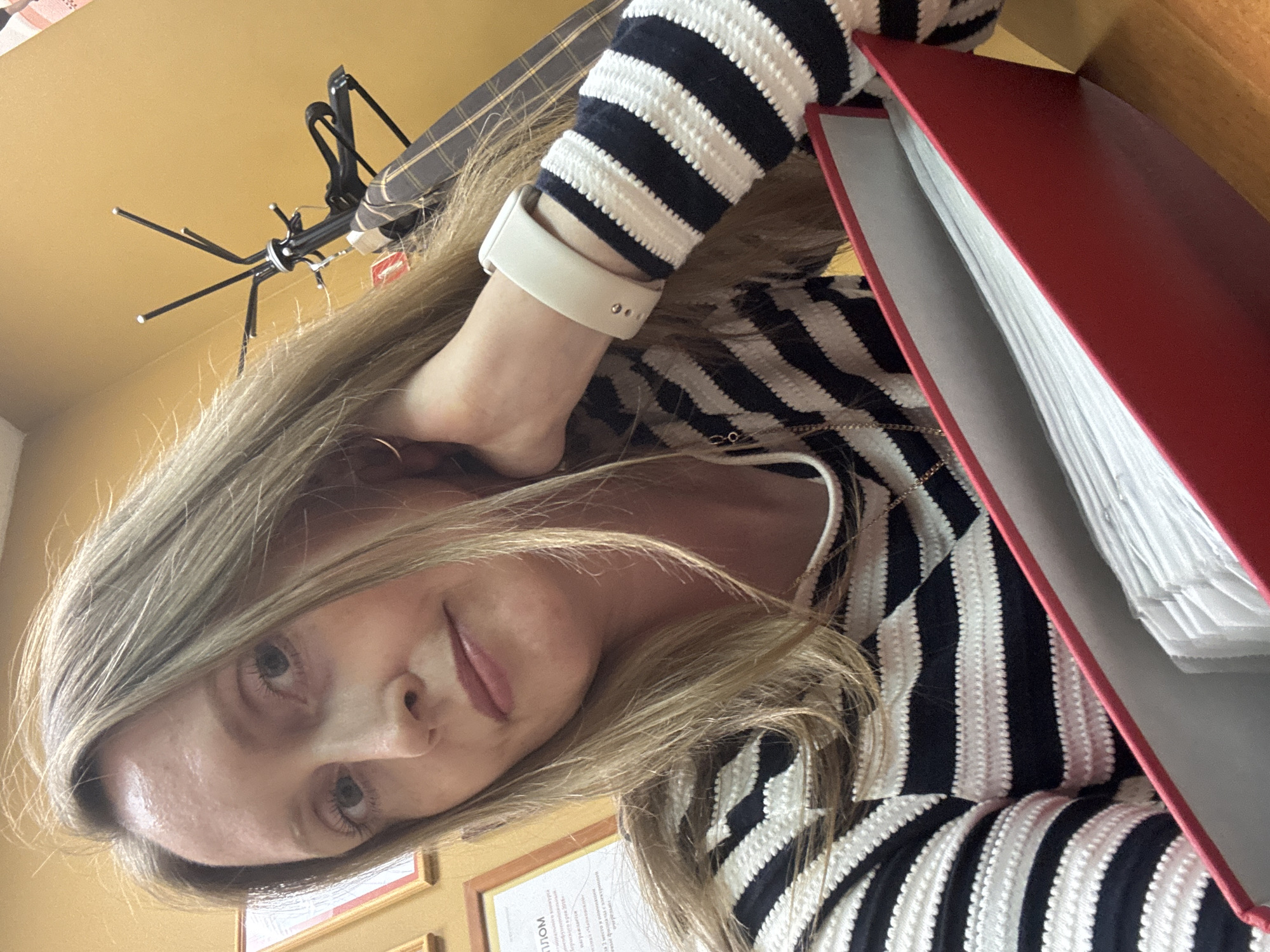 A selfie shows a woman with blonde hair and a white smartwatch wearing a black and white striped sweater. A red binder is to her right. Behind her is a wall with framed documents and what appears to be a clothes hanger. The setting is possibly an office or study. It's difficult to confirm the location as Chaikovsky, Russia, without additional visual or contextual clues.